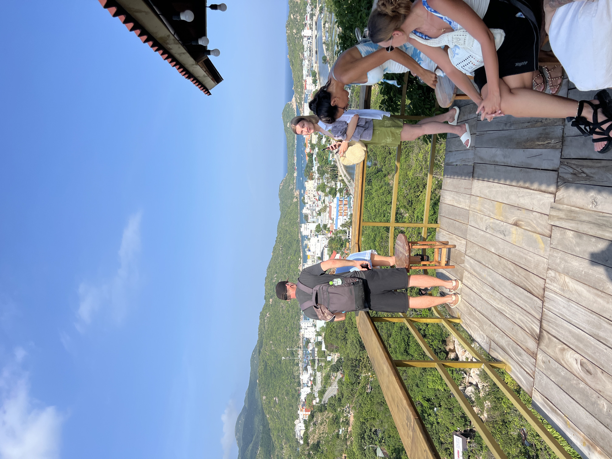 The image captures a panoramic view from a raised wooden deck overlooking a coastal town in Vĩnh Hải, Vietnam, on a clear, sunny day. Seven individuals are visible on the deck.
In the left foreground, a man, seen from behind, wears a black cap, dark grey t-shirt, black shorts, and tan sneakers, with a dark grey backpack containing a water bottle. He stands at the wooden railing, observing the vista. A small child in light clothing is partially visible next to him, also facing the view.
Towards the center, a blonde woman stands, holding a straw bag and a smartphone. She wears a light green skirt, a white open long-sleeved shirt, and white slide sandals, looking towards the left of the frame. To her right, a woman with dark hair tied up, wearing a blue and white patterned dress, leans forward over the railing. Further right, a woman with dark hair tied back, wearing sunglasses, a white top with blue accents, and black shorts, sits on a wooden stool with her hands clasped. She wears black strappy sandals with an ankle bracelet, with a white woven bag beside her. Partial views of two other individuals are discernible at the bottom right.
The wooden deck is constructed from weathered planks, featuring a sturdy wooden railing supported by golden-colored metal posts. In the upper right corner, a red corrugated roof with exposed wooden beams extends over the deck, from which several white lightbulbs hang.
The background reveals a vibrant blue sky with minimal white clouds. Below the deck, dense green foliage covers a hillside. The mid-ground showcases a compact coastal town composed of numerous white and light-colored buildings with red roofs, nestled between lush green mountains. A winding road traverses the town, and a bridge crosses a narrow waterway. The town opens into a calm, blue bay dotted with multiple small boats.