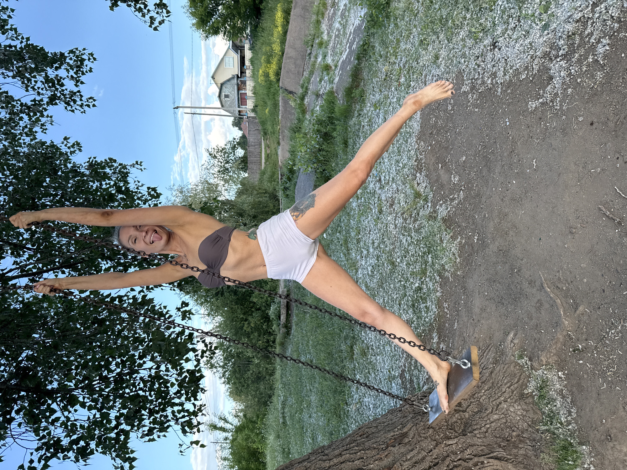 A woman with light-colored hair is hanging from a swing by her arms, with her legs spread in a wide split. She is wearing a brown bikini top and white shorts. Her tongue is sticking out and her eyes are closed. The swing is made of a wooden plank suspended by chains from a tree. The ground beneath the swing is covered with small white objects, possibly remnants of hail. In the background, there are trees, a rural house, and a bright blue sky with scattered clouds. The location is identified as the city of Chaikovsky, Russia.