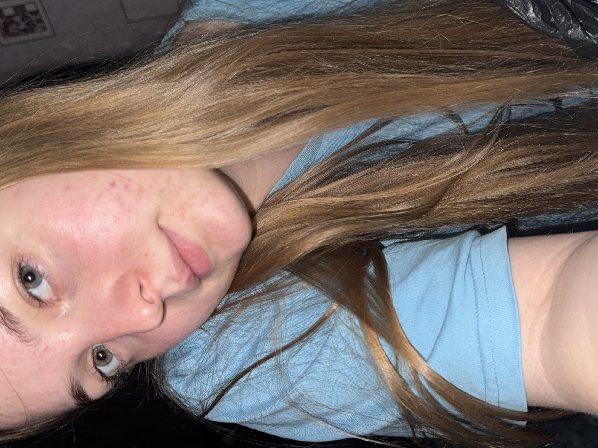 A person with light skin, light brown hair, and green eyes is lying down wearing a light blue t-shirt. The person's face is slightly tilted to the left and they are looking directly at the camera. The background is dark, with some indistinct items visible. There is no evidence to suggest that this photo was taken in Perm, Russia.