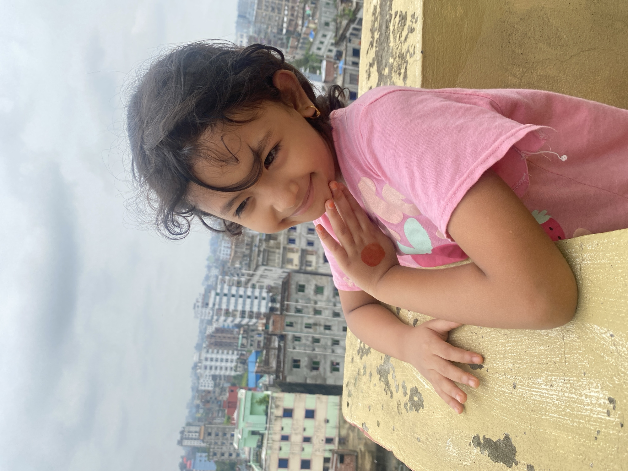 A young girl with curly dark hair and a pink t-shirt is posing on a rooftop overlooking a dense urban landscape. She has a bright smile and her left hand is raised to her chin, with a circular henna design visible on her hand. The background shows numerous buildings of varying heights, characteristic of the city of Dhaka, Bangladesh. The sky is overcast, suggesting a cloudy day. The girl is leaning against a textured, yellowish wall or railing.