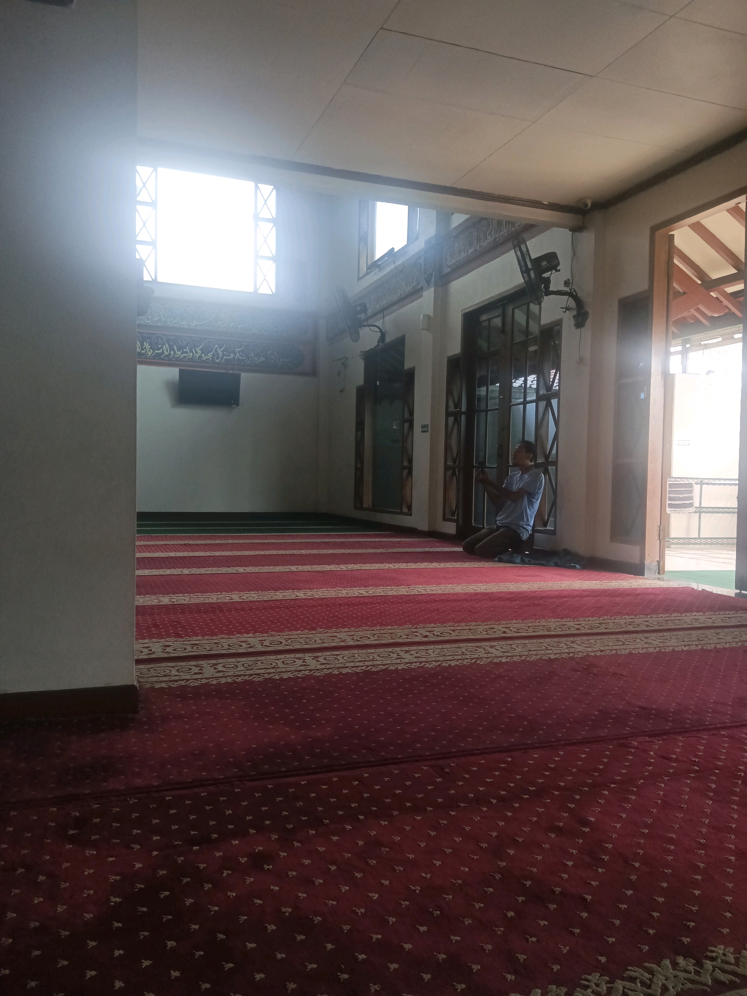 In a mosque in South Tangerang, Indonesia, a man kneels on a red carpet with a repeating pattern of small light-colored motifs. The carpet extends to the foreground and middle ground of the image, with rows of green carpet visible further in the distance. The man, wearing a blue t-shirt and dark pants, is facing a wall with Arabic calligraphy and has his hands clasped together in front of him. Behind him, there are large glass doors with dark frames and a ceiling fan mounted on the wall. Sunlight streams in through a large window in the background, illuminating the space. A black television screen is mounted on the wall to the left of the man. The overall scene depicts a moment of prayer within the mosque.