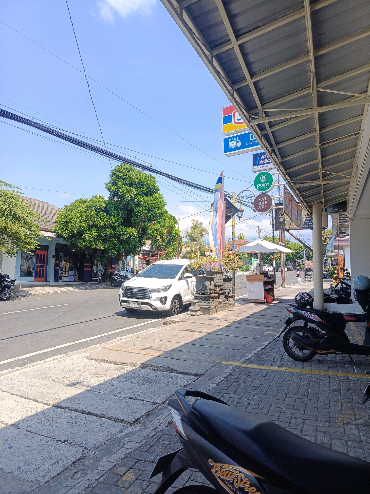 The image depicts a street scene in the city of Kepolorejo, Indonesia, on a clear, sunny day. A white Toyota Innova car is parked on the right side of the road, facing away from the camera. Adjacent to the road is a sidewalk paved with grey bricks and concrete slabs, shaded by the awning of a building. Several motorcycles are parked on the sidewalk. The building features a corrugated metal roof and is decorated with signs, including one for "Point Coffee."
Across the street, on the left side, is a row of buildings with blue and orange facades, one with an orange door. A large, leafy green tree stands prominently in front of one of these buildings. Overhead, a network of black electrical wires is strung across the sky. A few people and more motorcycles are visible in the distance. The overall atmosphere is that of a typical commercial street in a tropical urban environment.