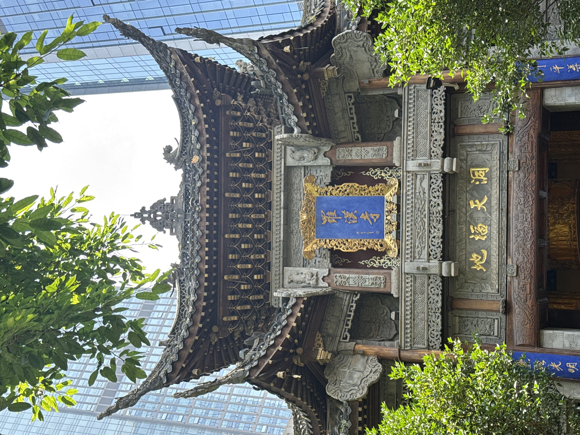The image captures a richly adorned traditional Chinese architectural structure, likely a pavilion or gate, under bright daylight. The building features an elaborate roof with distinctive upturned eaves and intricate wooden and stone carvings throughout its facade, columns, and beams. Gilded decorative elements enhance the traditional craftsmanship.
A prominent blue plaque with golden Chinese characters "觀音閣" (Guanyin Ge), translating to "Guanyin Pavilion," hangs centrally. To its right, a vertical blue banner reads "大慈悲殿" (Dacibei Dian), meaning "Great Hall of Compassion." Further to the right, on a door, another plaque displays "迴廊" (Huí Láng), or "Corridor."
Lush green foliage partially frames the structure, with sunlit leaves adding a natural element to the scene. In the background, contrasting sharply with the traditional architecture, a modern glass skyscraper with a grid pattern rises under a clear, bright sky. This juxtaposition highlights the integration of historical sites within a contemporary urban environment, characteristic of the Yuzhong District, China.
The scene is illuminated by bright, direct sunlight, suggesting a clear day during late morning or early afternoon. No people or specific activities are visible, emphasizing the architectural beauty and its setting. The image evokes a sense of cultural heritage preserved amidst modern development.