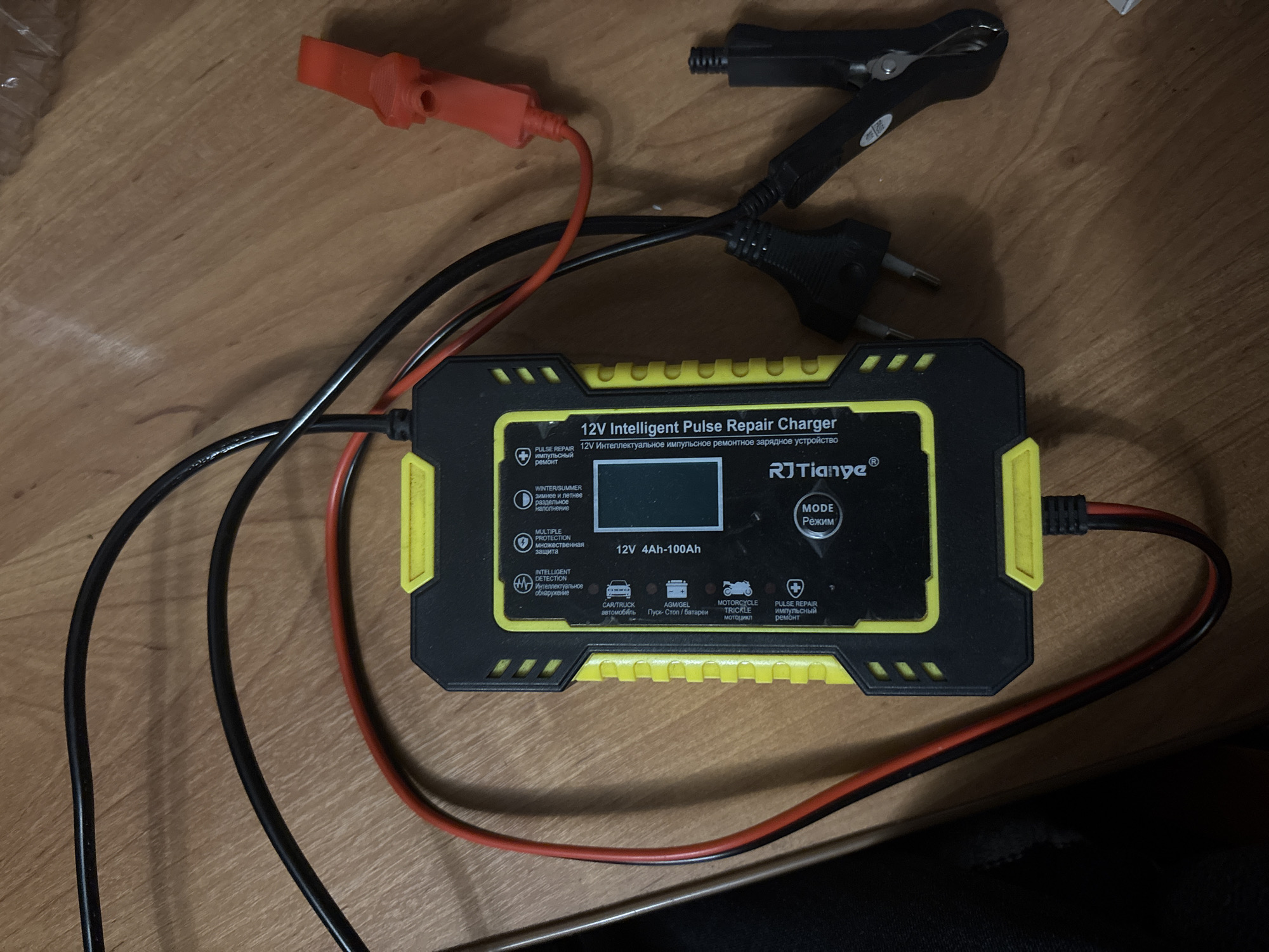 The image shows a 12V Intelligent Pulse Repair Charger, a device for maintaining or repairing vehicle batteries. It is a black and yellow device with various modes for different types of batteries (car, truck, motorcycle, AGM/GEL). The charger has red and black leads with clamps for connecting to a battery. The charger has a European plug. The location is indiscernible, it is just a wooden table. There is no information in the image to suggest the location is Zagatuy, Russia.