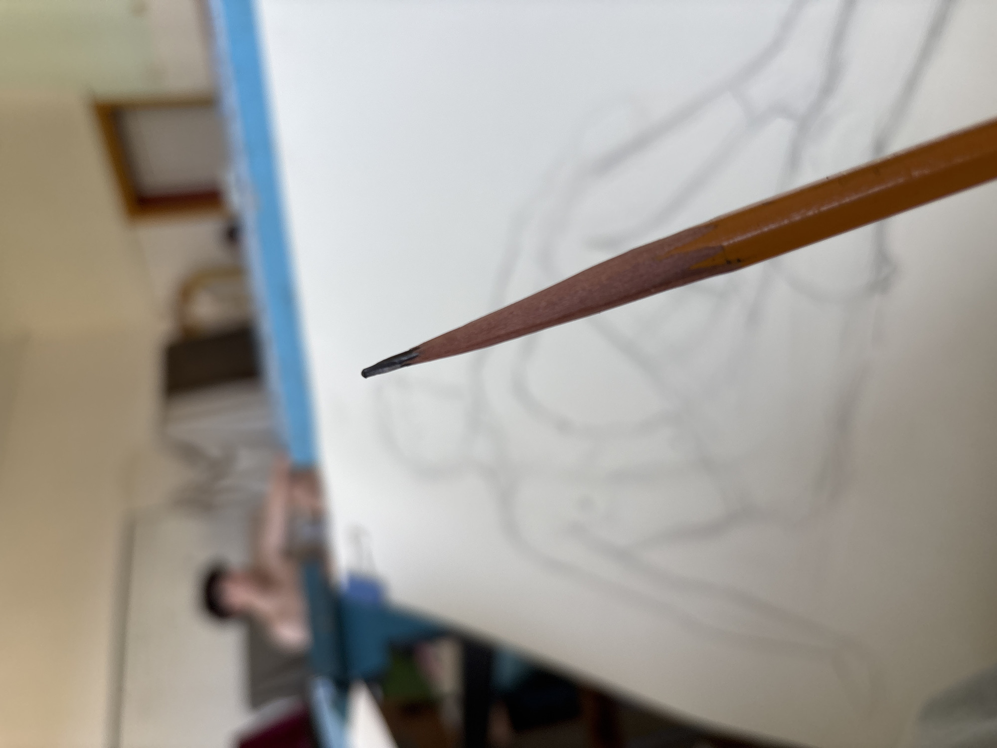 The image captures an indoor artistic setting in Troyan, Bulgaria, with a strong focus on a sharpened wooden pencil in the foreground. The pencil, dark brown with a distinct black graphite tip, is held at an angle, poised as if actively being used for drawing.
In the blurred background, a large white sheet of paper or a drawing board is visible, leaning against what appears to be an easel or a drafting table with a noticeable blue border. On this white surface, a faint graphite sketch is discernible, suggesting a figure study or an intricate organic form. Behind the drawing setup, a highly blurred individual can be seen, likely the artist, seated or standing, engaged in the creative process.
Further back, the room elements are indistinct but include blurred shapes that could be a window or a framed opening, allowing natural light to enter. The overall atmosphere suggests a moment of concentration and artistic endeavor. There is no visible text in the image.
