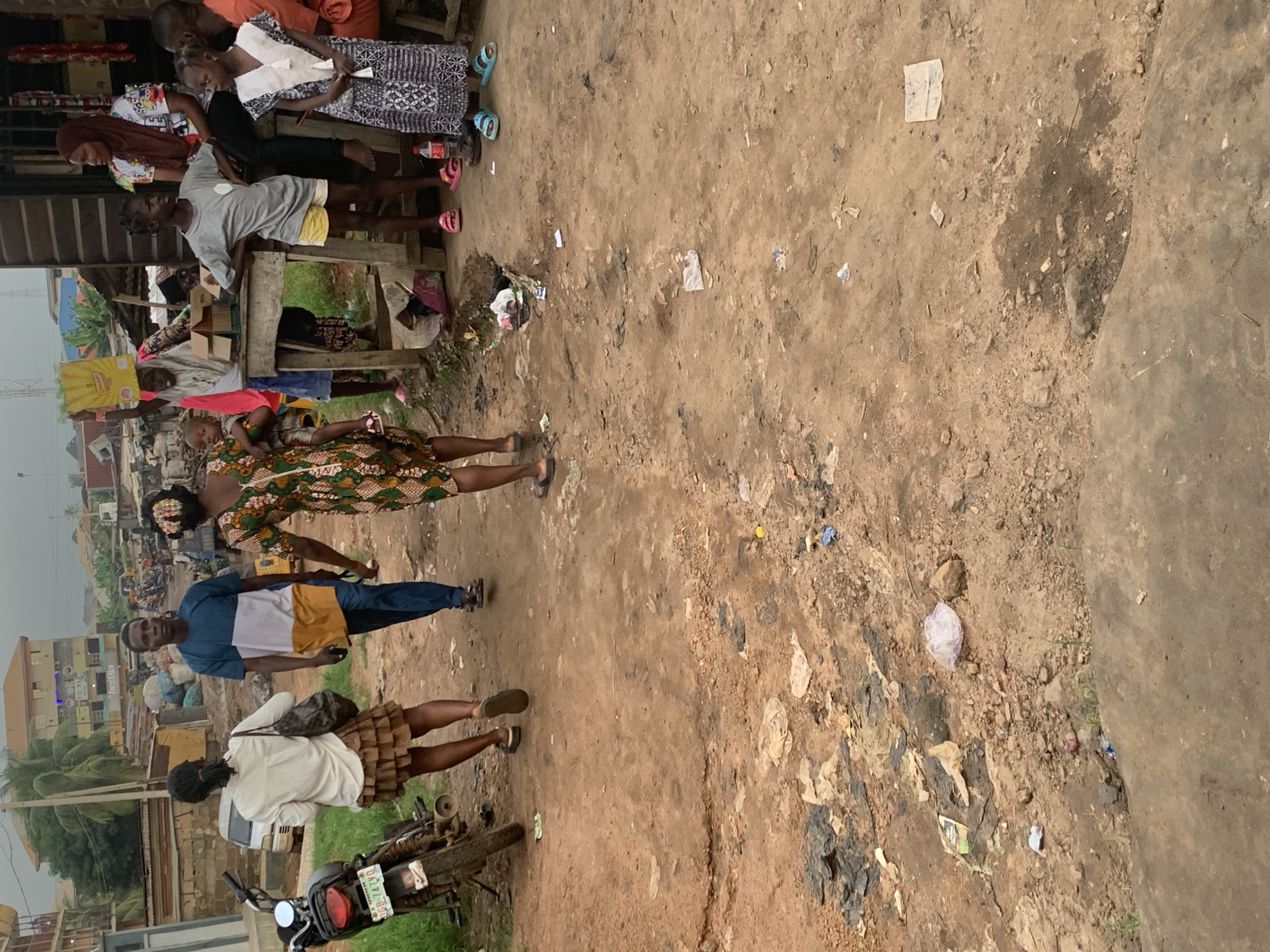 This media file depicts a street scene in Shagamu, Nigeria. The primary subjects are several people, including adults and children, engaged in everyday activities. One woman is walking, wearing a patterned dress and a headscarf. Another person, a man, is standing and looking towards the camera, dressed in a blue and yellow shirt and blue trousers. Further down, a woman in a white top and a brown skirt is standing near a motorcycle, which is parked on the dirt ground. In the background, there are buildings, including a multi-story structure, and glimpses of vegetation. The ground is unpaved and appears to be a dirt road with scattered debris, suggesting an outdoor market or a less developed urban area.
The overall impression is that of a casual day in a bustling Nigerian town. The lighting suggests it is daytime, possibly overcast given the diffused light. The presence of people in a relaxed stance and the general atmosphere indicate normal daily life. There is no specific event clearly visible, rather a snapshot of people going about their business.
Notable details include the varied clothing styles, from traditional patterns to more contemporary attire. The presence of a motorcycle hints at local transportation methods. The condition of the ground, with its dirt and scattered refuse, provides a strong visual cue about the environment. While no specific emotions are overtly displayed, the scene is not one of distress or urgency. The focus is on the candid depiction of life in this location. No visible text is clearly legible within the frame.