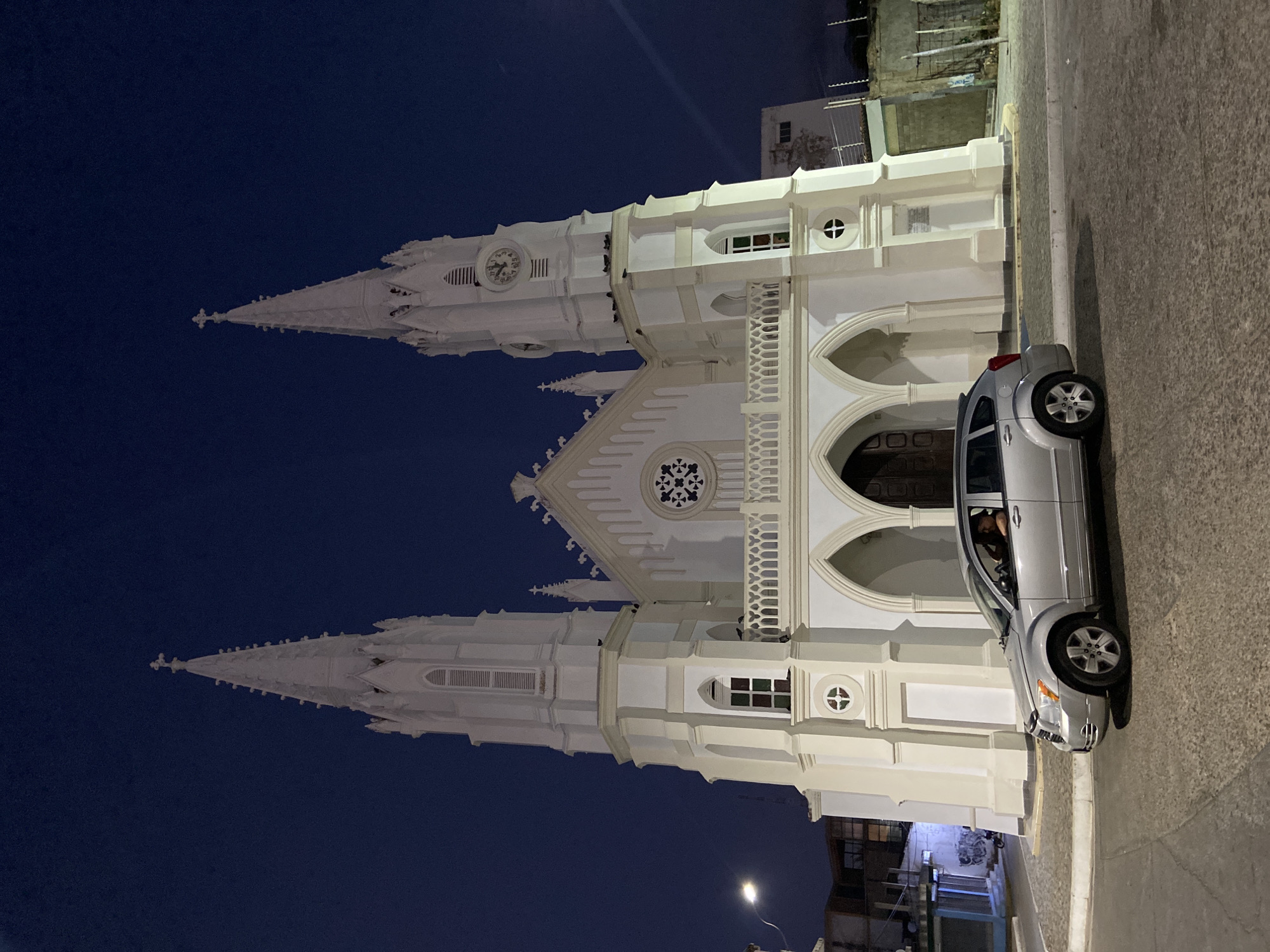 At night, a white church with two towers and spires stands next to a street in Juan Griego, Venezuela. A silver vehicle is parked on the street next to the church. The night sky is dark blue. Buildings are visible in the background. A street light illuminates the buildings in the background.