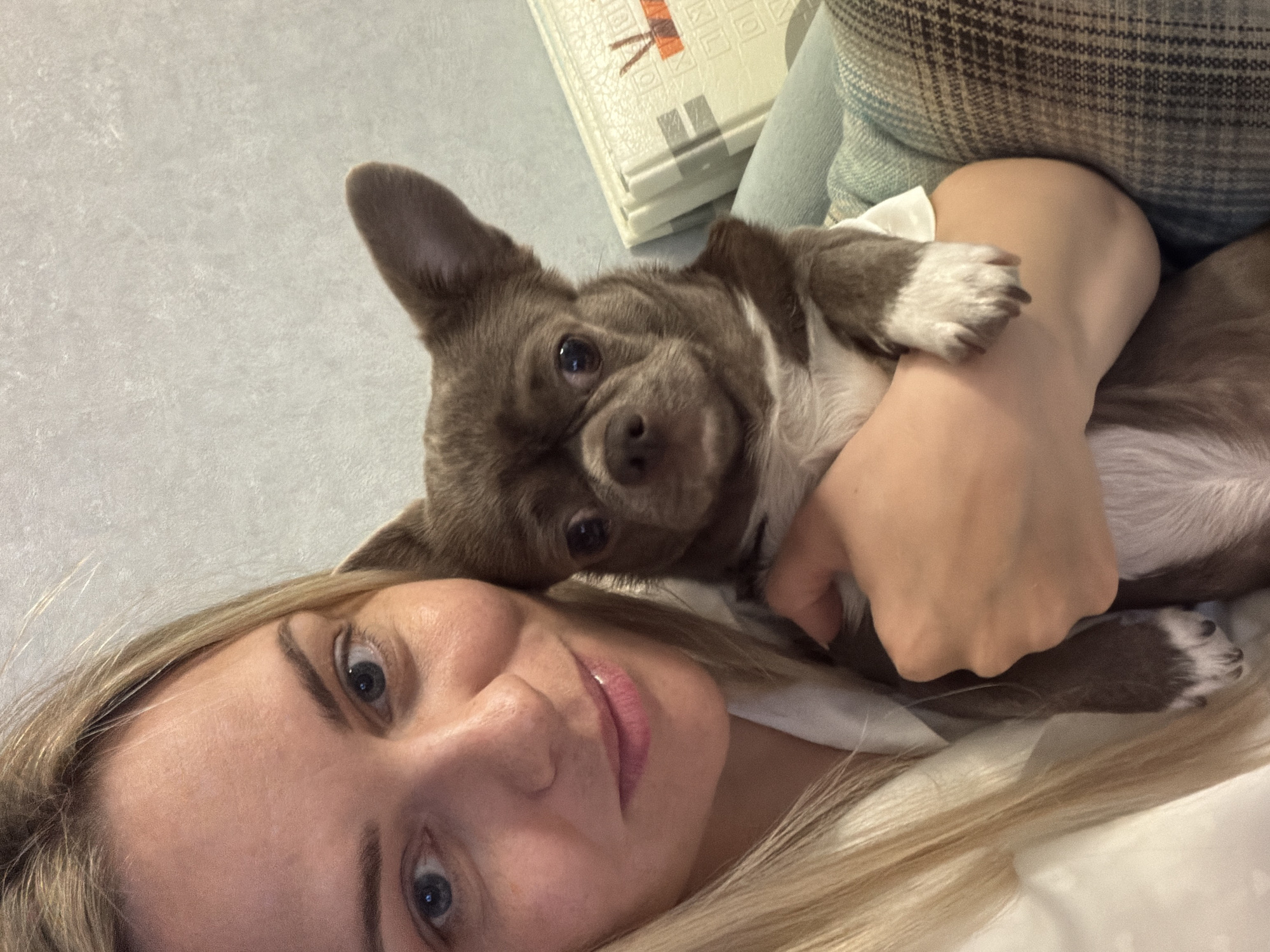 A close-up selfie-style image captures a woman and a small dog sharing a tender moment indoors. The woman, with long blonde hair, bright blue eyes, and light pink lipstick, lies on her side in the lower left of the frame, looking directly at the camera with a soft, content expression. Her face occupies a significant portion of the foreground. Nestled closely beside her, in the upper right of the frame, is a small, short-haired dog, likely a Chihuahua. The dog has brown-grey fur with distinctive white markings on its chest and paws, and large, upright ears. Its dark eyes are also focused on the camera, giving a curious and attentive gaze. The woman's arm is visible, gently holding the dog close to her body. The setting appears to be a cozy indoor space, possibly a bed or sofa, indicated by soft textures and a patterned pillow or blanket behind them. In the background, a light-colored, textured surface is visible, which may be a floor or playmat, featuring faint, indistinct patterns and partial shapes that are not clearly decipherable as text. The overall atmosphere is one of warmth, affection, and companionship. The location context is Votkinsk, Russia.