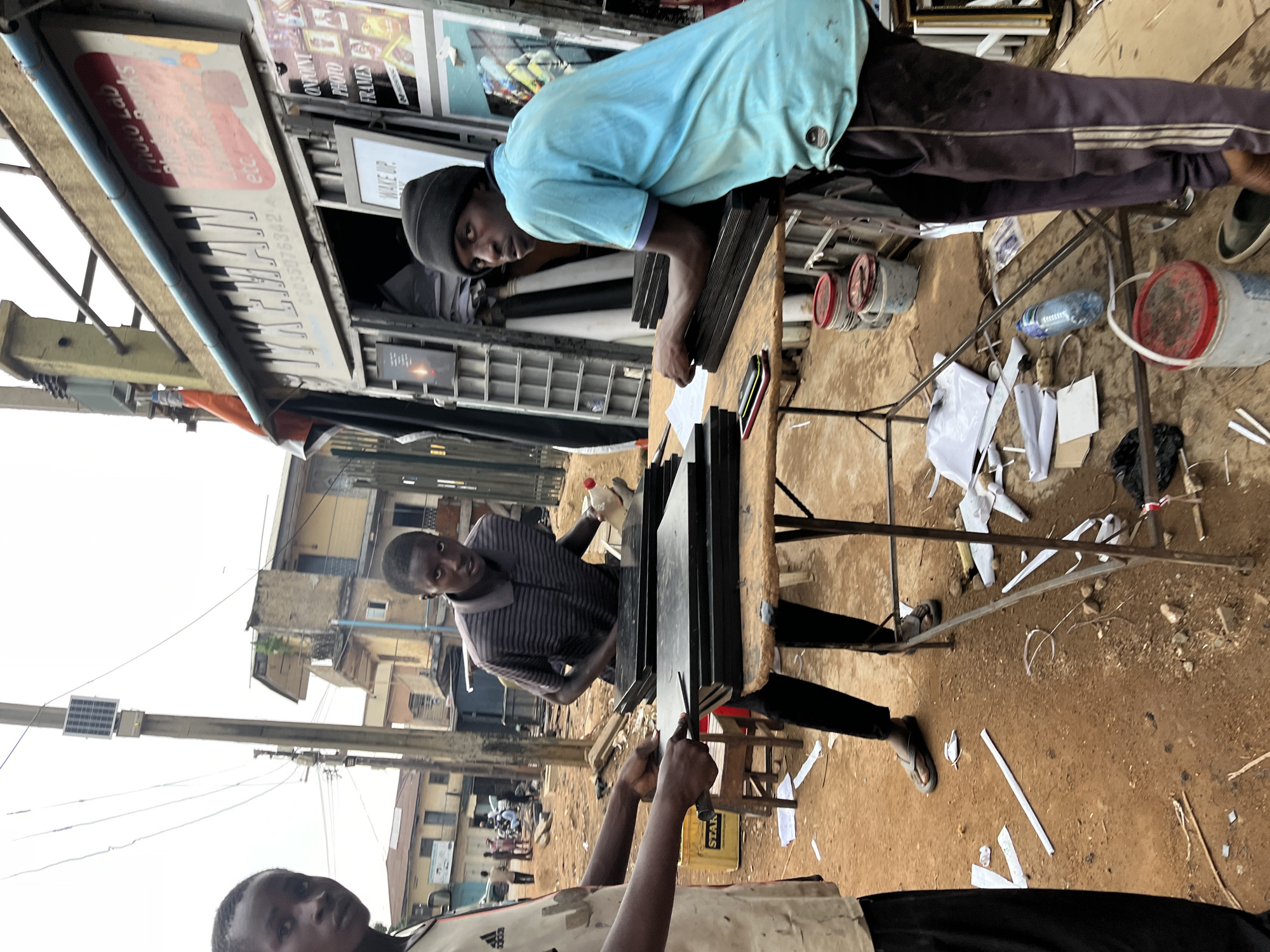 The image depicts an outdoor scene in Jos, Nigeria, likely a commercial area with a small shop. In the foreground, three young males are visible. The person on the left is partially visible, wearing a light-colored shirt with "adidas" branding. The central figure, dressed in a striped shirt and dark pants, appears to be handling a stack of dark, flat objects on a rustic wooden table. To the right, a third male, wearing a light blue t-shirt and dark pants with white stripes, leans over the table, looking towards the camera. He is also near the stack of objects.
The shop in the background has a sign with text including "TYKEMAN" and "PHOTO LAB." Displayed on the shop's facade are various framed pictures and posters, suggesting it is a photography or framing business. The ground is unpaved and appears to be a dirt or gravel surface. Scattered on the ground are papers, debris, and several buckets, some with lids. Electrical wires and a utility pole are also prominent in the background, indicating an urban or semi-urban setting. The overall environment suggests a busy, working area.