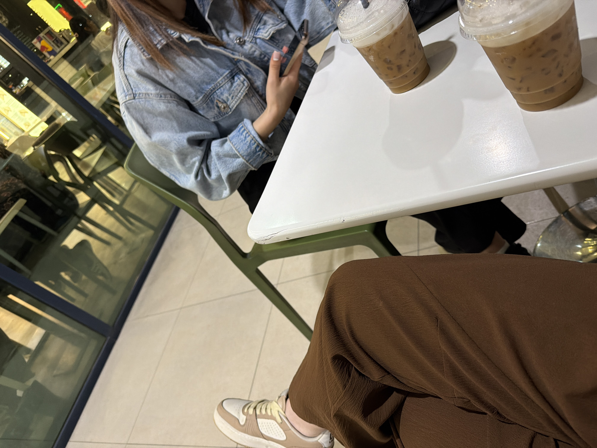 The image shows two people sitting at a white table in what appears to be a cafe or similar establishment. One person is wearing a light denim jacket and holding a smartphone, while the other is wearing brown pants and tan and white shoes. Two iced coffee drinks are on the table. The location appears to be Yerevan, Armenia based on the setting and storefronts visible through the windows. The scene depicts a casual social interaction over drinks.