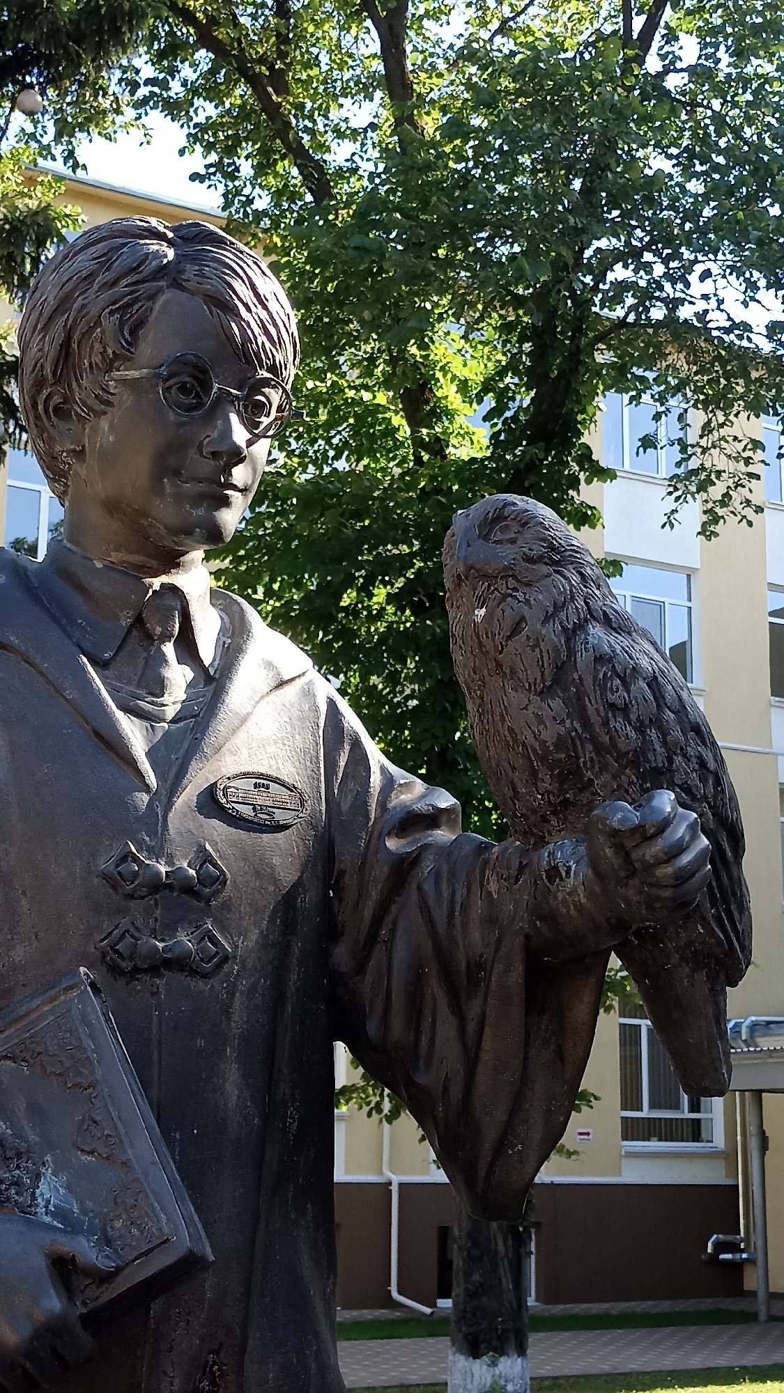 The image depicts a bronze statue of a young person wearing glasses and a robe, holding a book in their left hand. An owl perches on the statue's right arm. The statue is situated outdoors, with a building and trees in the background. This statue is located at the Tiraspol State University in Tiraspol, Moldova, dedicated to the university's founder, Grigori Stanchev.