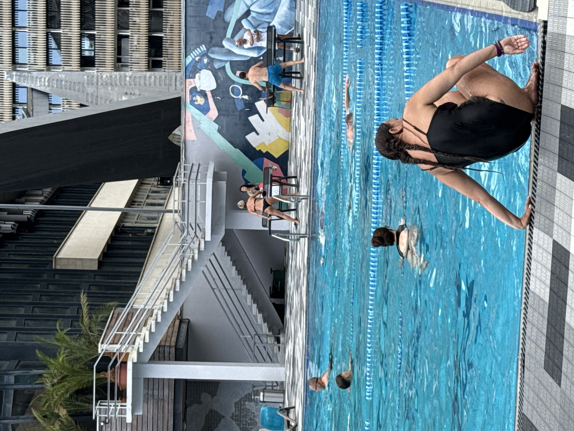 The image depicts an outdoor swimming pool with several people engaged in various activities. In the foreground, a woman with braided dark hair and wearing a black swimsuit is seated on the edge of the pool, facing away from the camera. Several other individuals are visible swimming in the pool, with lane markers dividing the water.
In the background, there is a large mural on a wall with abstract shapes and figures. Adjacent to the pool, there are tables, potentially for games like ping pong, with people interacting around them. Behind the pool area, a multi-story building with a modern architectural design is visible. The scene suggests a public recreational facility, possibly a swimming complex located in or near Массандровский поселковый совет, Ukraine.