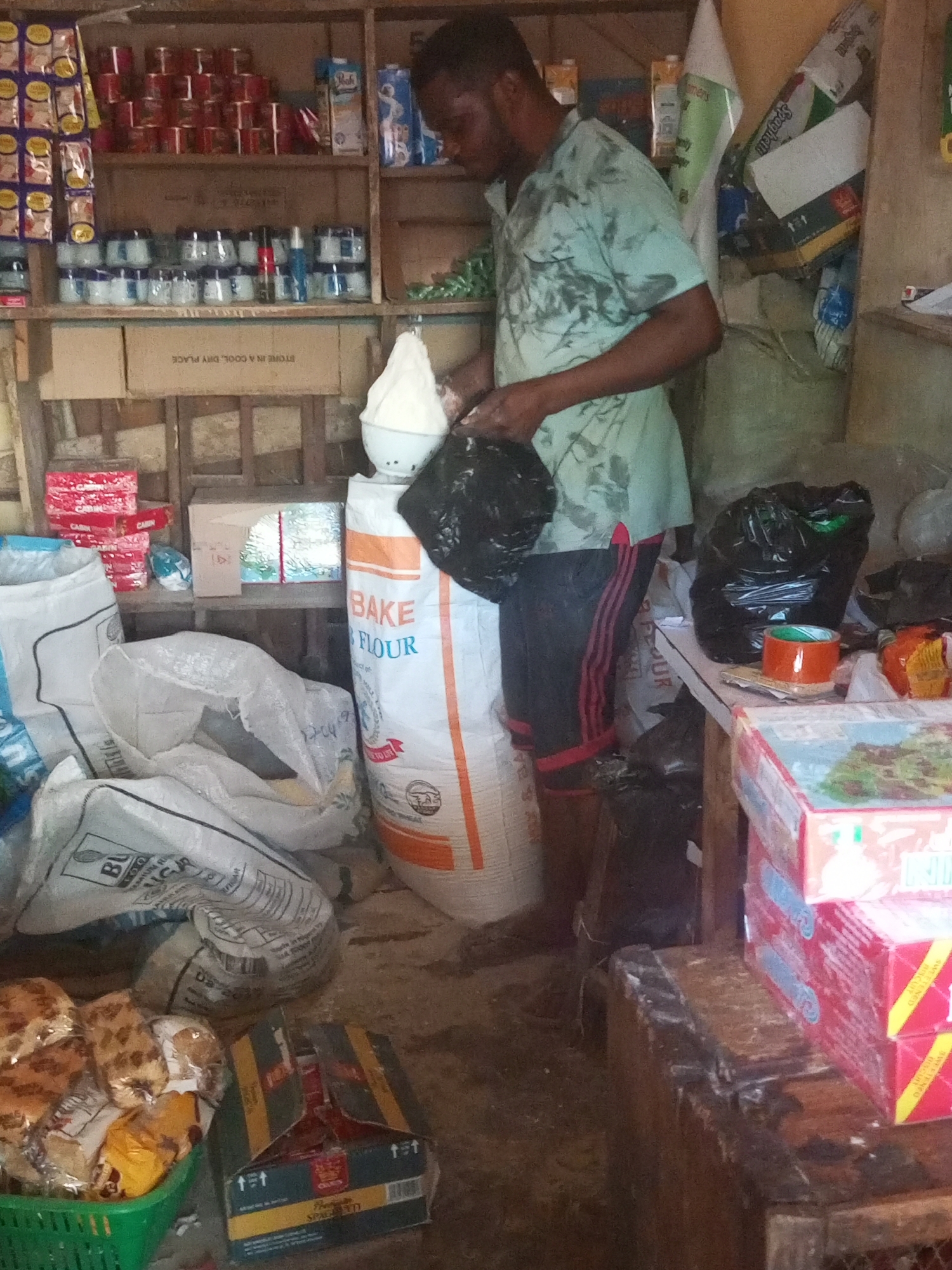 A man stands inside a small store located in Sabon Tasha, Nigeria. He wears a light green t-shirt and dark shorts with red stripes down the sides. He is in the process of filling a black plastic bag with white powder, likely flour, scooped from a white bowl. A large bag of "BAKE FLOUR" is visible near him. The store's shelves are stocked with various items, including canned goods, jars, and boxed products. Bags of flour and other goods are scattered on the floor. The overall setting appears to be a modest, local retail establishment.