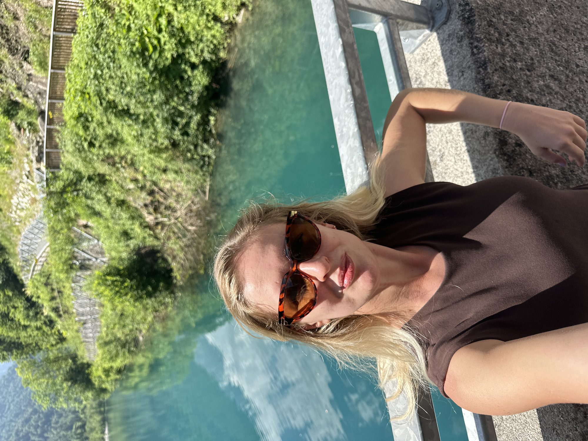 A woman with blonde hair and sunglasses takes a selfie in front of a vibrant turquoise lake in Kaprun, Austria. She is wearing a brown t-shirt and a thin pink bracelet. The lake is surrounded by lush green trees and hills, with a pathway visible along the embankment. A metal railing runs along the foreground of the image. The water reflects the sky and clouds, creating a serene and picturesque scene.