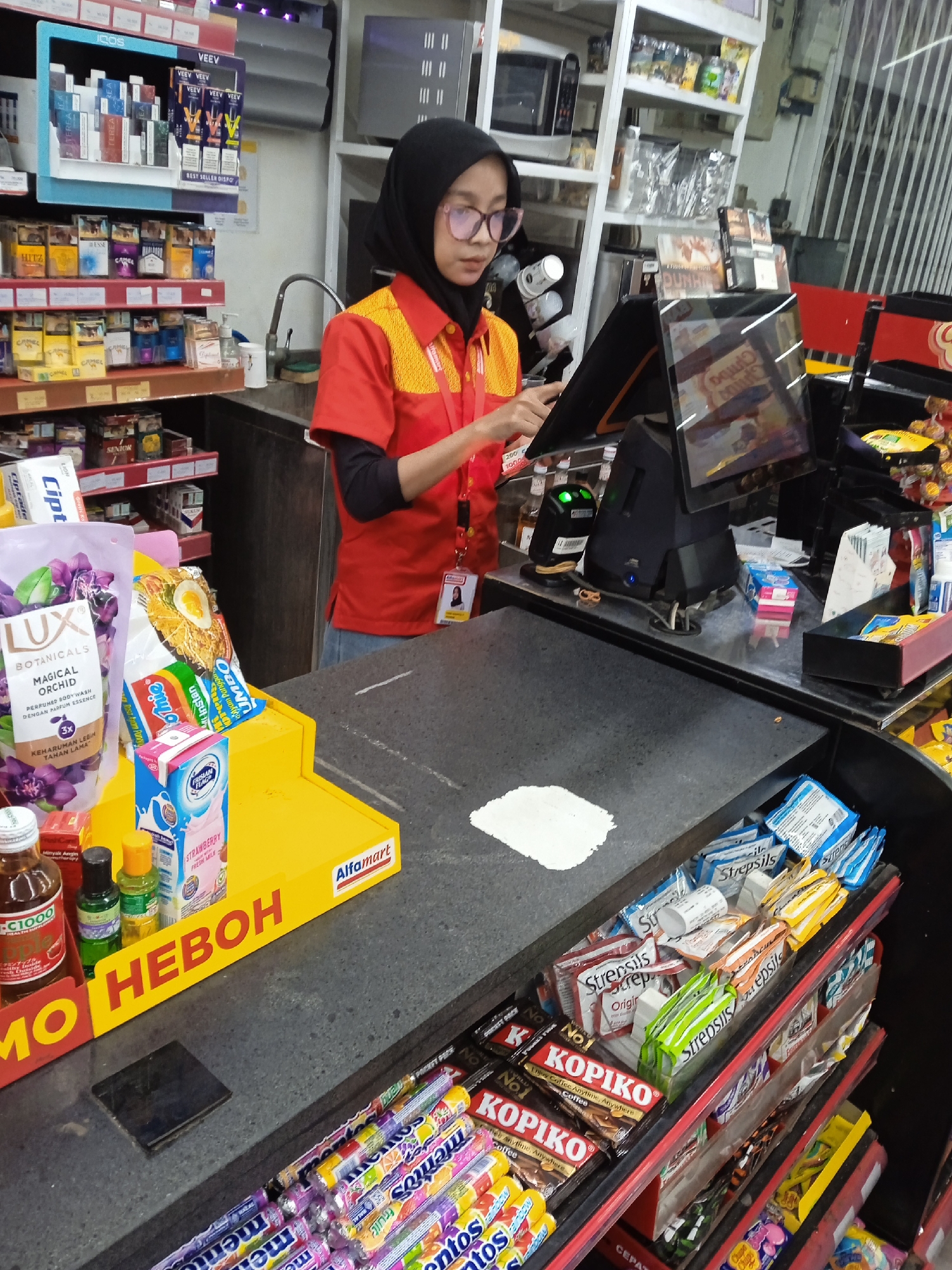 The image depicts a female employee at the checkout counter of an Alfamart convenience store, likely located in Karangtengah, Indonesia. She is wearing a black hijab, pink-rimmed glasses, and a red and yellow Alfamart uniform shirt, with blue jeans or an apron visible below. She is focused on and operating a touchscreen cash register, with a second display screen facing the customer.
The store counter is well-stocked with various impulse purchase items. On a bright yellow display stand, the word "HEBOH" is prominently displayed in red, alongside the "Alfamart" logo. Products visible on this stand include a large bottle of "LUX BOTANICALS MAGICAL ORCHID PERFUMED BODY WASH," a packet of Indomie instant noodles (specifically "Ayam Panggang UMBO"), and a carton of "STRAWBERRY FRESH MILK." Smaller bottles, such as "C1000" and "Minyak Angin," are also on display. Beneath the counter, shelves hold numerous small packages of "mentos," "Kopiko" coffee candies ("No.1 Coffee Anytime, Anywhere"), and various flavors of "Strepsils" lozenges, including "Original."
Behind the employee, shelves are fully stocked with a range of tobacco and vape products, including brands like "VEEV" (advertised as "BEST SELLER DISPO"), "IQOS," "Marlboro," "Camel," "Esse," "Hitz," and "Dunhill." A hand sanitizer dispenser is also visible near a small sink. The indoor lighting suggests typical store operating hours. The employee appears to be diligently performing her duties, demonstrating focus rather than any strong emotion. Other partially visible texts include "Ciptadent" and "SENIOR."