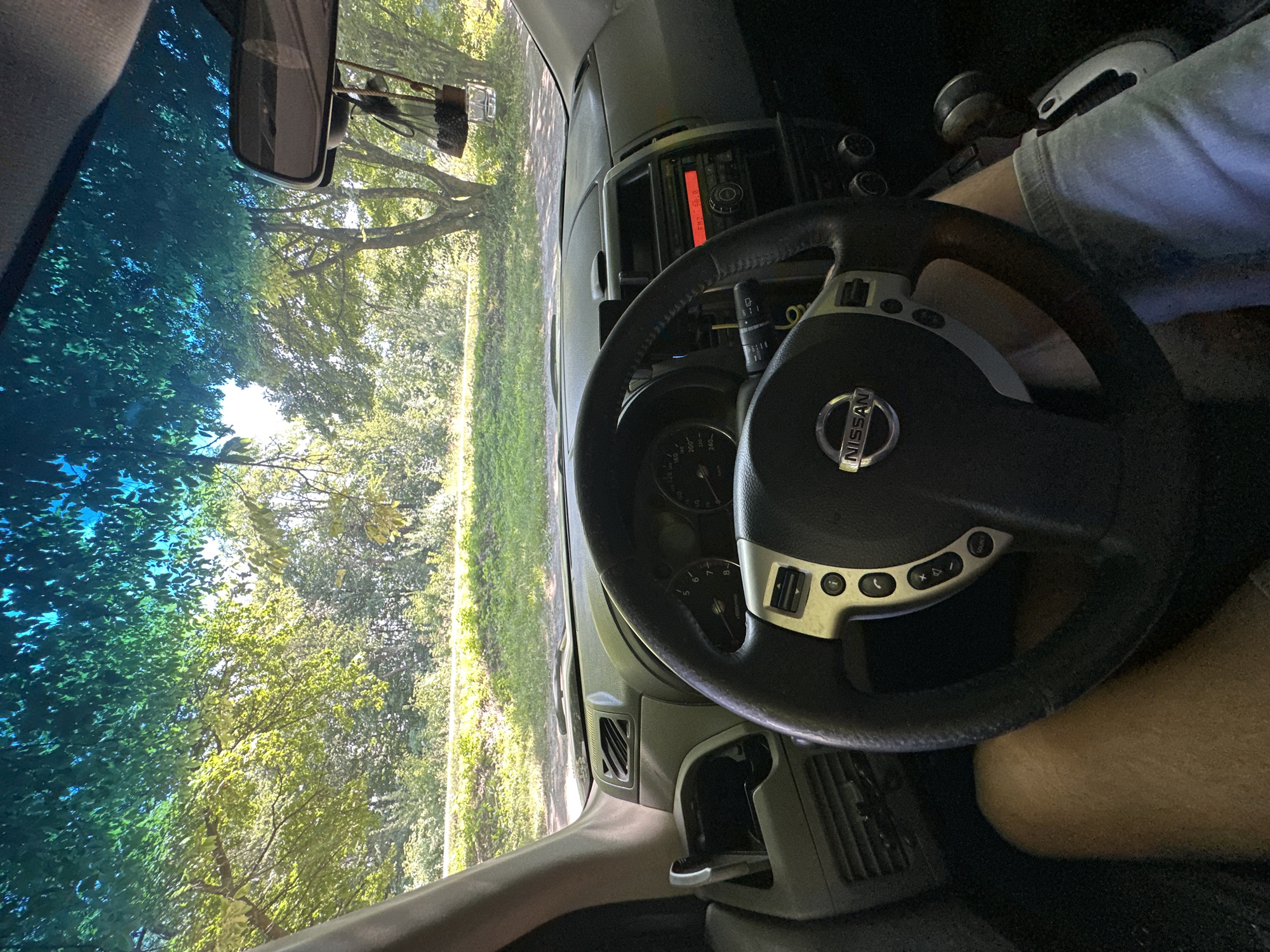 The image shows the interior of a car, looking forward through the windshield. The steering wheel, dashboard, and part of the driver's leg are visible in the foreground. The car appears to be parked or stopped, with trees and greenery visible outside the windshield. The scene is well-lit by natural sunlight filtering through the leaves and windshield. There are no people visible other than the driver's leg. The overall impression is one of a peaceful, natural setting, likely a rural area or park.