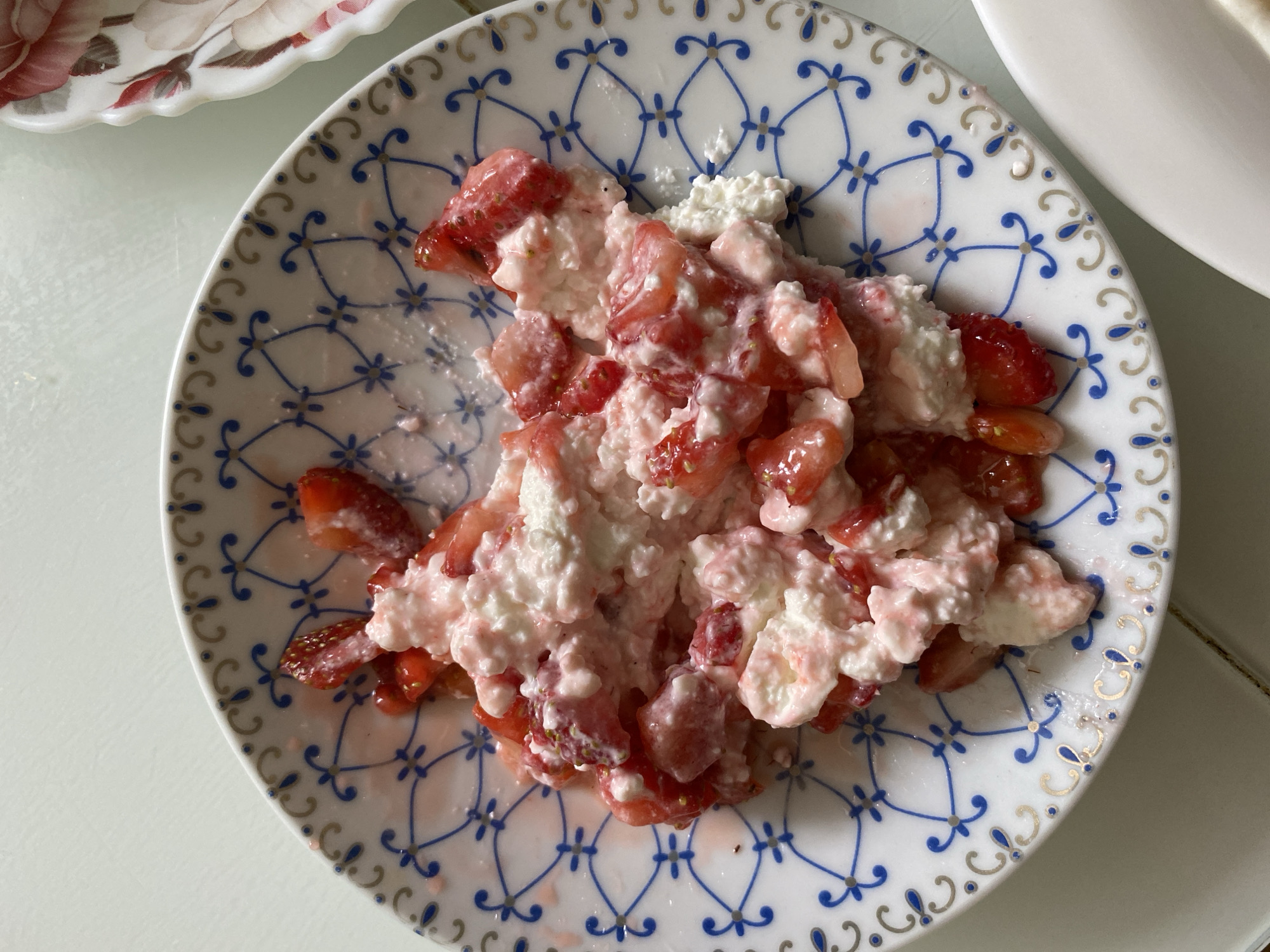 A close-up, top-down view shows a plate filled with cottage cheese and sliced strawberries. The plate has a white background with a blue and gold decorative pattern around the rim. The food is piled in the center of the plate, with some of the pink strawberry juice staining the white parts of the plate. The background is a light-colored surface, likely a table. No other objects or people are clearly visible. The scene appears to be indoors, in Votkinsk, Russia.