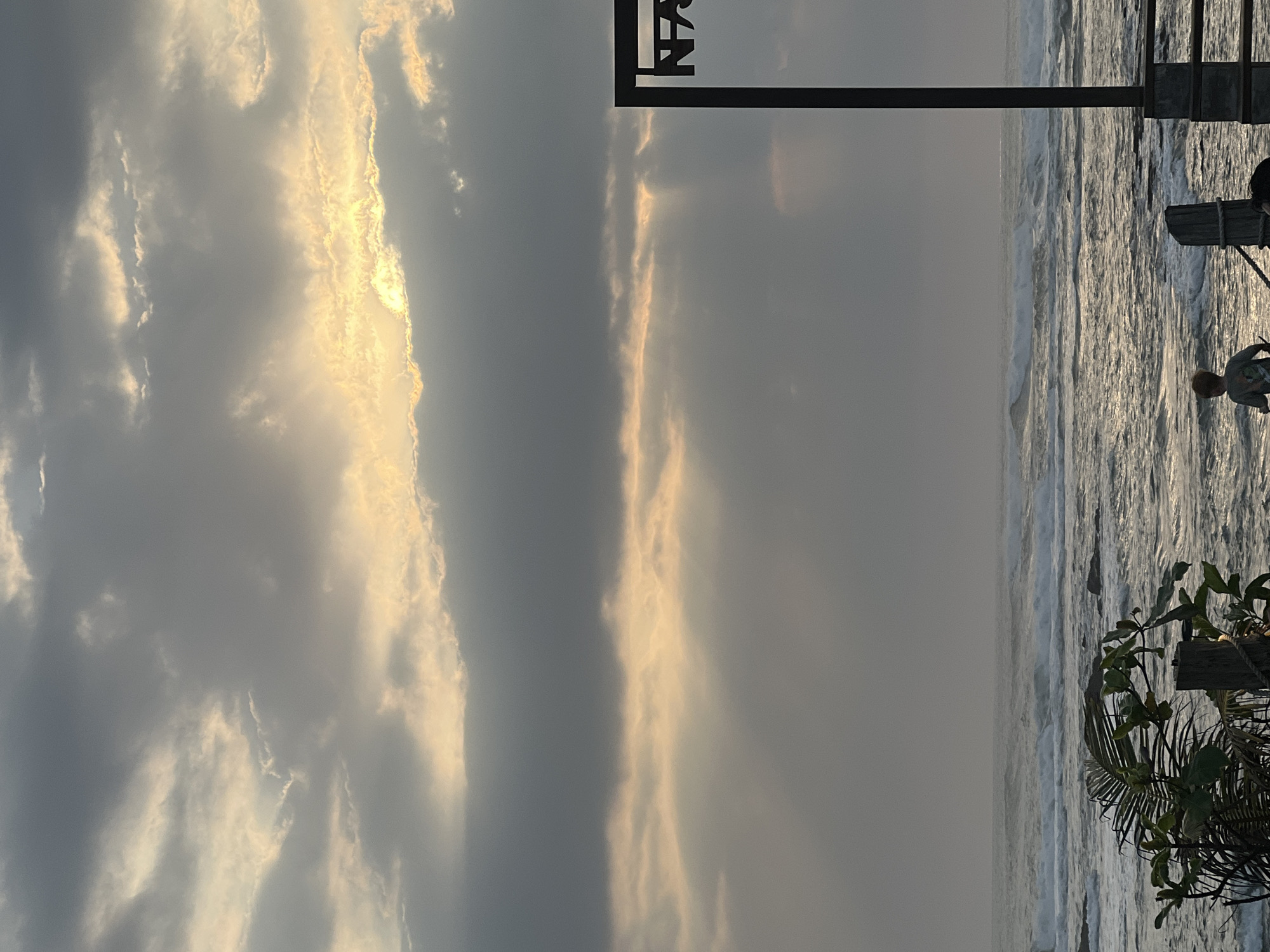 The image depicts a cloudy sky over a choppy ocean. Sunlight breaks through the clouds, creating rays of light that illuminate the water. In the foreground, a young boy is walking near the shore, facing the ocean. To the right, a dark, angular structure with the letters "NA" visible on it stands near the water. Vegetation, including palm fronds and branches, is also present in the foreground. The scene appears to be a beach in the city of Mengwi, Indonesia.