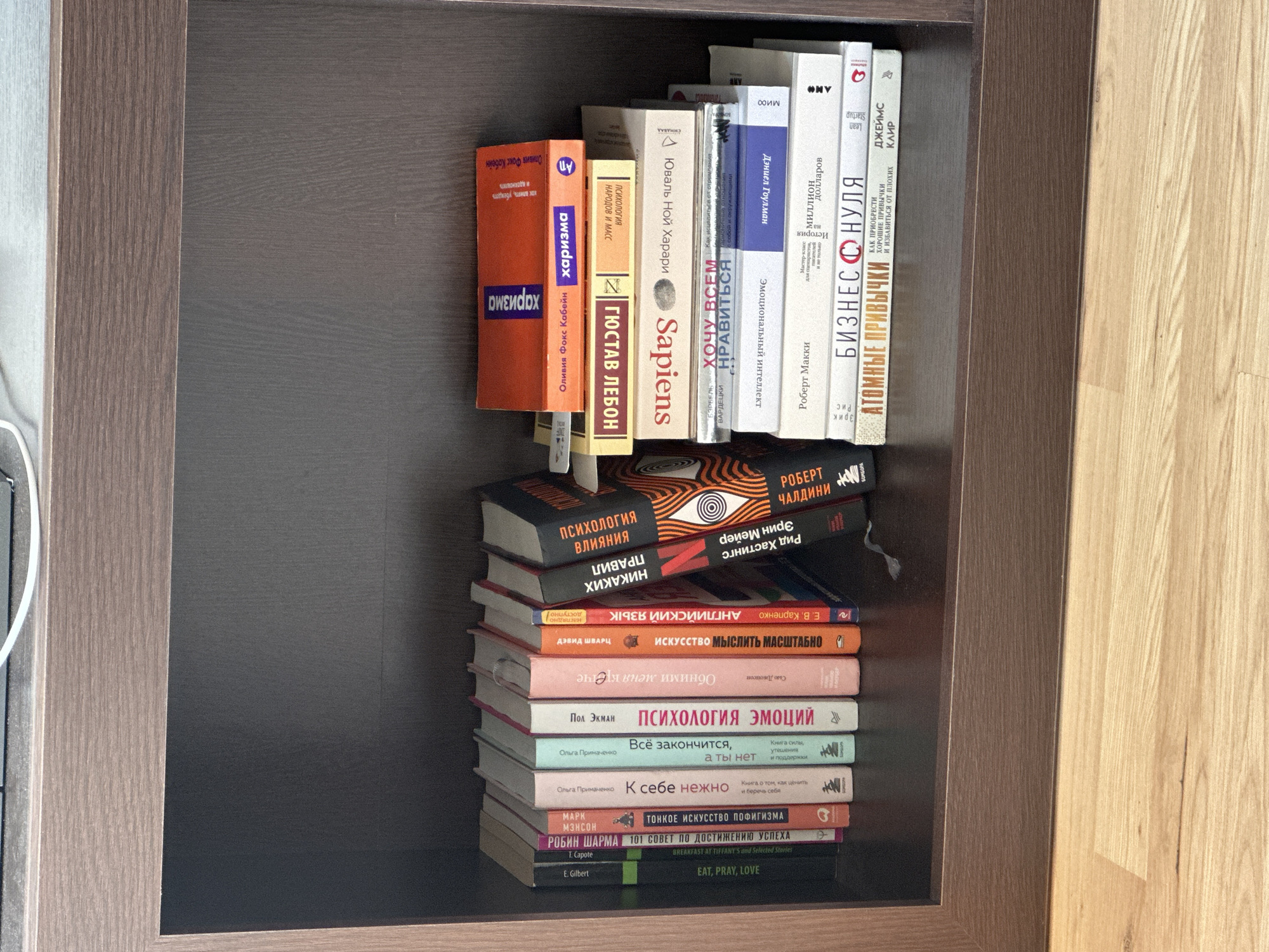 The image shows a bookshelf filled with books in what appears to be a private residence, likely a home. The books are arranged in two stacks. The top stack consists of books standing upright, oriented as if ready to be read. The books are mostly in Russian, with titles visible such as "Sapiens," "Atomic Habits," and books on psychology and emotional intelligence. The bottom stack has books piled on top of each other, titles including "Eat, Pray, Love," and "The Subtle Art of Not Giving a F*ck." The bookshelf itself is dark brown with lighter wood trim on the side, suggesting it is a built-in unit. The books vary in size and color, giving a cluttered, yet well-used, aesthetic to the shelf. Based only on the image content, there is no way to confirm the location in Boreti, Montenegro, or any other particular location.