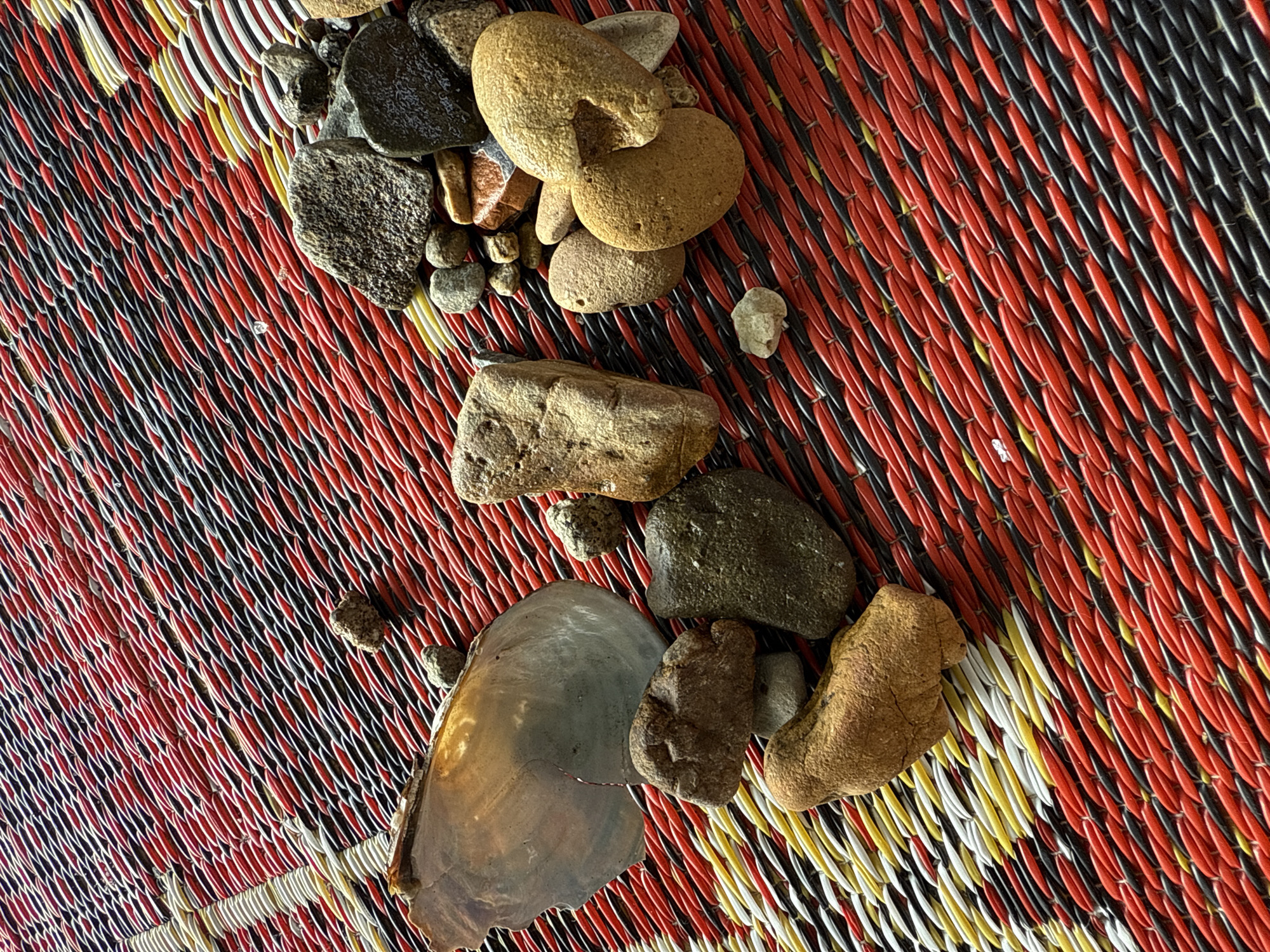 A close-up, high-angle shot shows a collection of natural objects on a woven mat with red, black, and yellow stripes. The mat's texture is intricate, with each strand appearing tightly twisted. Scattered across the mat are various stones, ranging in size, shape, and color from dark gray to light brown. Some stones are smooth and rounded, while others are more angular and rough. In addition to the stones, a large, iridescent mussel shell fragment is visible on the left side of the frame. The lighting suggests it is daytime, casting shadows that highlight the textures of the objects and the mat. The scene appears to be laid out for display or examination, possibly in an outdoor setting in Parapat, Indonesia, given the natural elements. No people are present in the image.