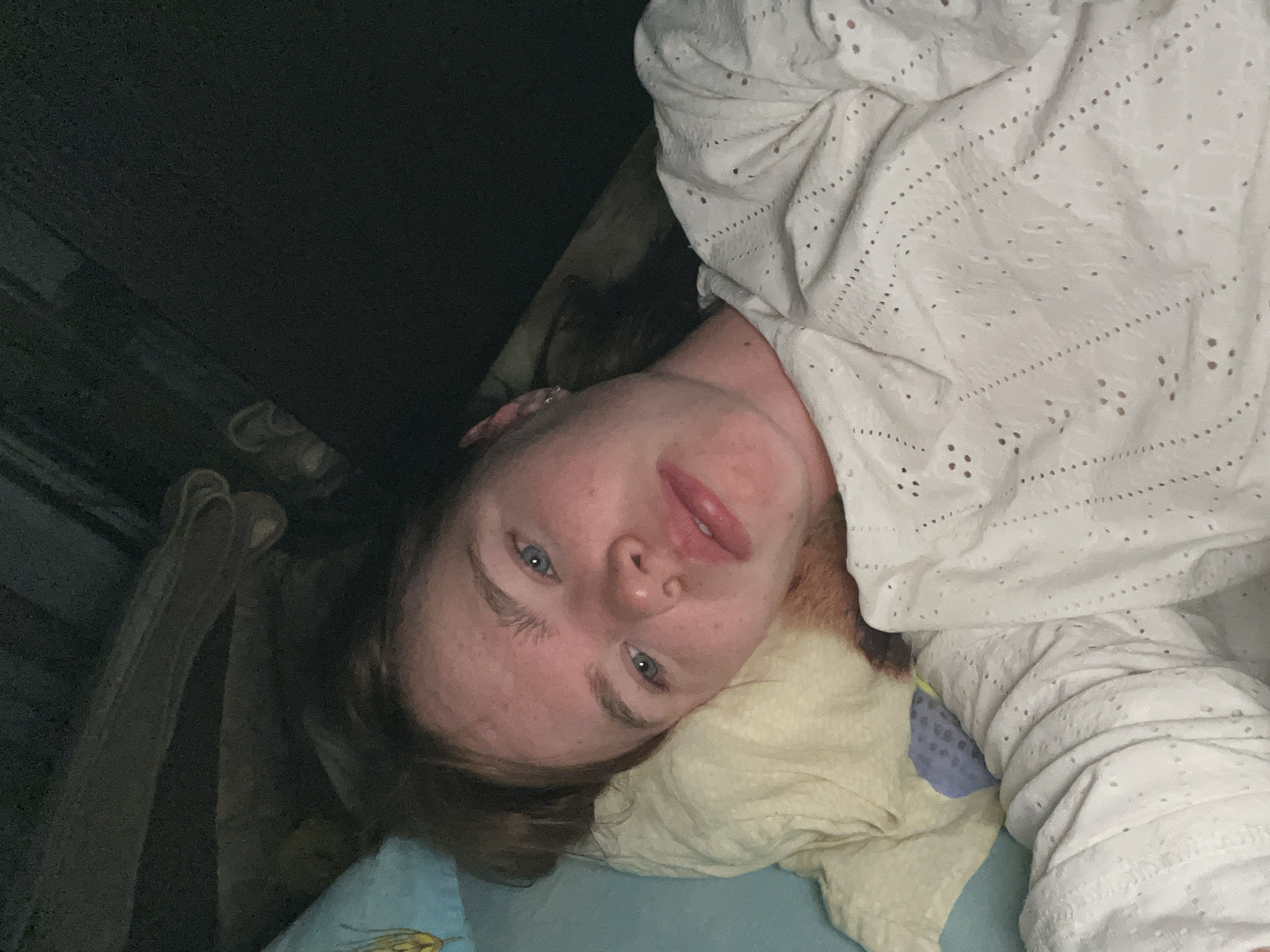 A person is lying on a bed with a light blue sheet and a yellow pillow. The person is covered with a white eyelet-patterned blanket. The person has fair skin, light eyes, and light brown hair. The location cannot be determined from the image.