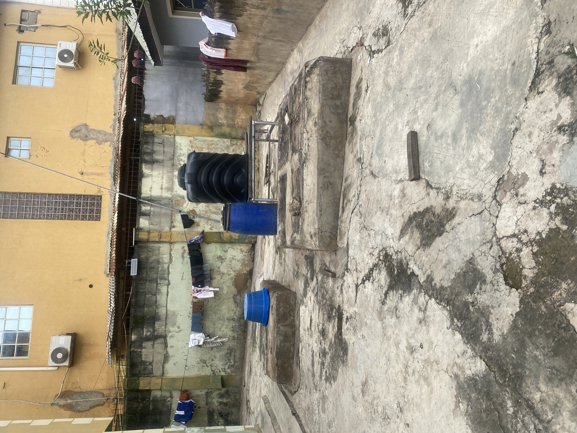 The image depicts an outdoor rooftop or courtyard area in Abeokuta, Nigeria, characterized by weathered concrete surfaces and a surrounding wall. A yellow building with multiple windows and an air conditioning unit is visible in the background. Laundry hangs on a clothesline stretched across the space, with various garments including shirts, shorts, and undergarments. Two large, dark blue water tanks are present, one notably tall and ribbed. In the foreground, there are two raised, rectangular concrete platforms, one of which has a bright blue plastic tub placed on top. Other objects scattered on the concrete include a piece of wood and some debris. The environment appears utilitarian and somewhat neglected, with visible cracks and staining on the concrete.