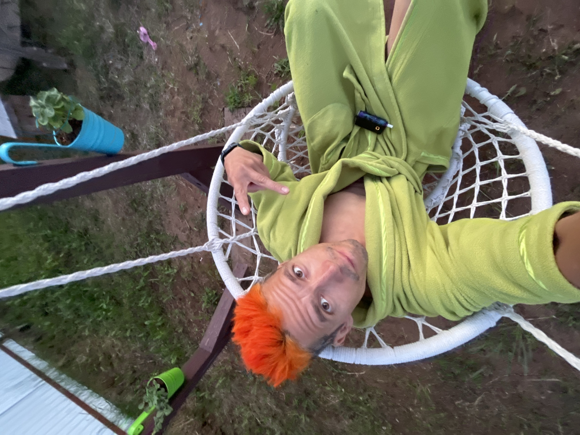 An overhead shot captures a man with striking orange, spiky hair and a light green, fleece-like robe, reclining in a white macrame hanging chair. He looks directly at the camera with wide eyes and a subtle smirk, holding his right hand up in a "V" or peace sign gesture. A black and purple vape pen is visible tucked into the fabric of his robe near his chest.
The scene is outdoors, likely a backyard or garden in Chaikovsky, Russia, with dry grass and dirt ground visible around and beneath the chair. To the left, a light blue plastic pot with a green plant hangs from a wooden support. Another smaller green plastic pot with a plant is seen on the ground to the right. White ropes forming the chair's suspension are prominent, and a thicker white rope stretches horizontally across the lower part of the frame, attached to a wooden beam.
The time of day appears to be daytime, possibly late afternoon, with soft, diffused lighting and no harsh shadows, suggesting an overcast sky or indirect light. The overall mood is casual and relaxed, with a touch of quirkiness from the subject's vibrant hair and direct engagement with the camera. No visible text is discernable in the image.