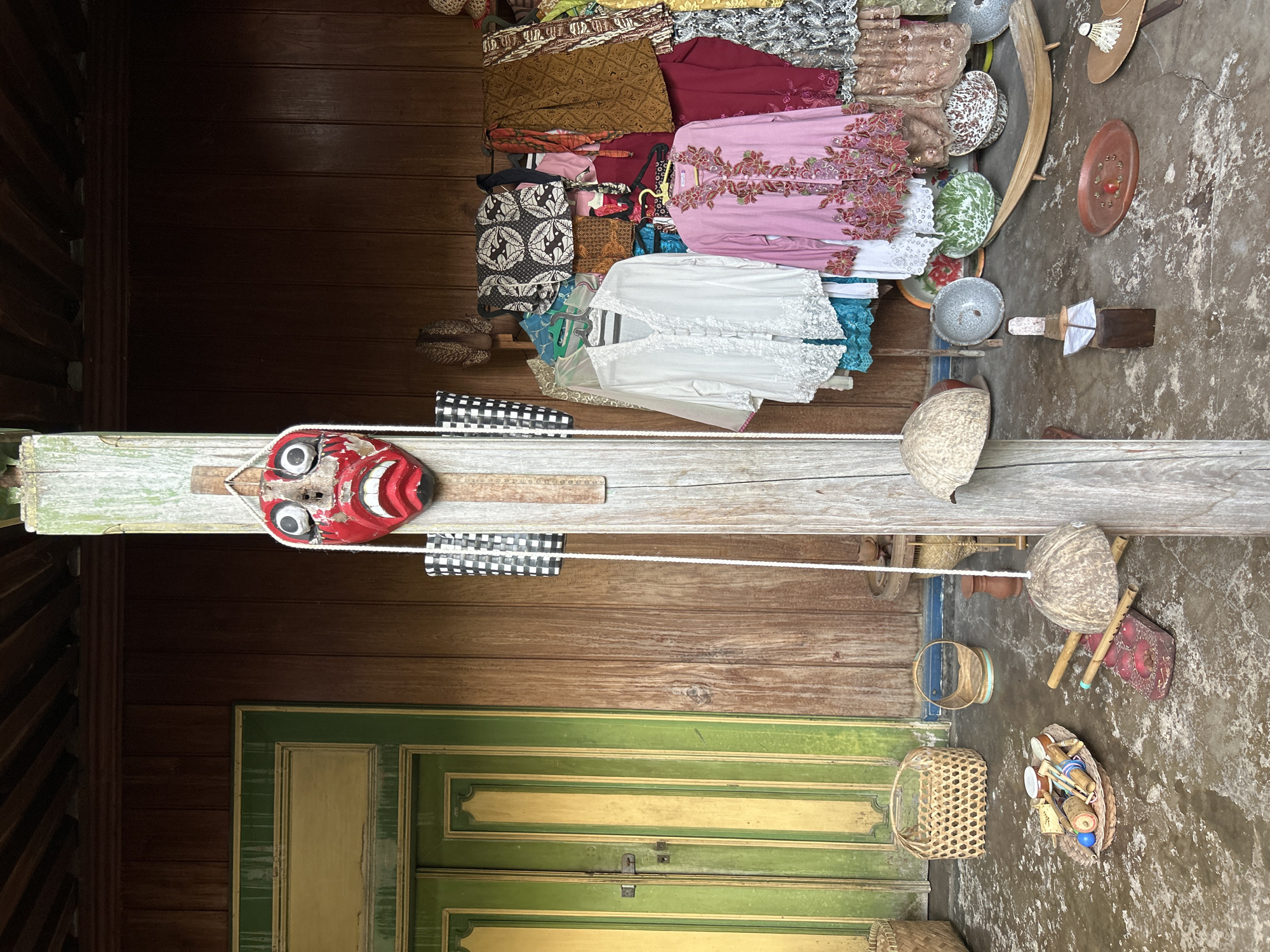 The image depicts a collection of traditional Indonesian crafts and clothing displayed on wooden surfaces. The setting appears to be an outdoor market or shop in Banguntapan, Indonesia, given the wooden architecture and the concrete ground with scattered debris. The scene is illuminated by natural light, suggesting it is daytime.
Prominently featured is a vibrant red and white wooden mask with large, expressive eyes, hanging from a wooden beam. Below the mask, a rope is strung across, with a black and white checkered fabric attached. To the left of the mask, part of a weathered wooden door with green and yellow painted panels is visible.
Behind the mask, a variety of colorful fabrics, possibly batik or traditional Indonesian garments, are neatly folded and displayed. These include intricately patterned cloths in shades of brown, maroon, pink, and white, some adorned with embroidery. Several woven baskets of different sizes and materials are also scattered around the scene. One basket contains small wooden items, possibly toys or decorative pieces. Other objects include metal bowls, a coconut shell, and what appear to be bamboo pipes.
The overall impression is one of a cultural display, showcasing the artistry and traditions of the region. The items are arranged in a somewhat rustic manner, suggesting an authentic, local setting. There is no visible text in the image.