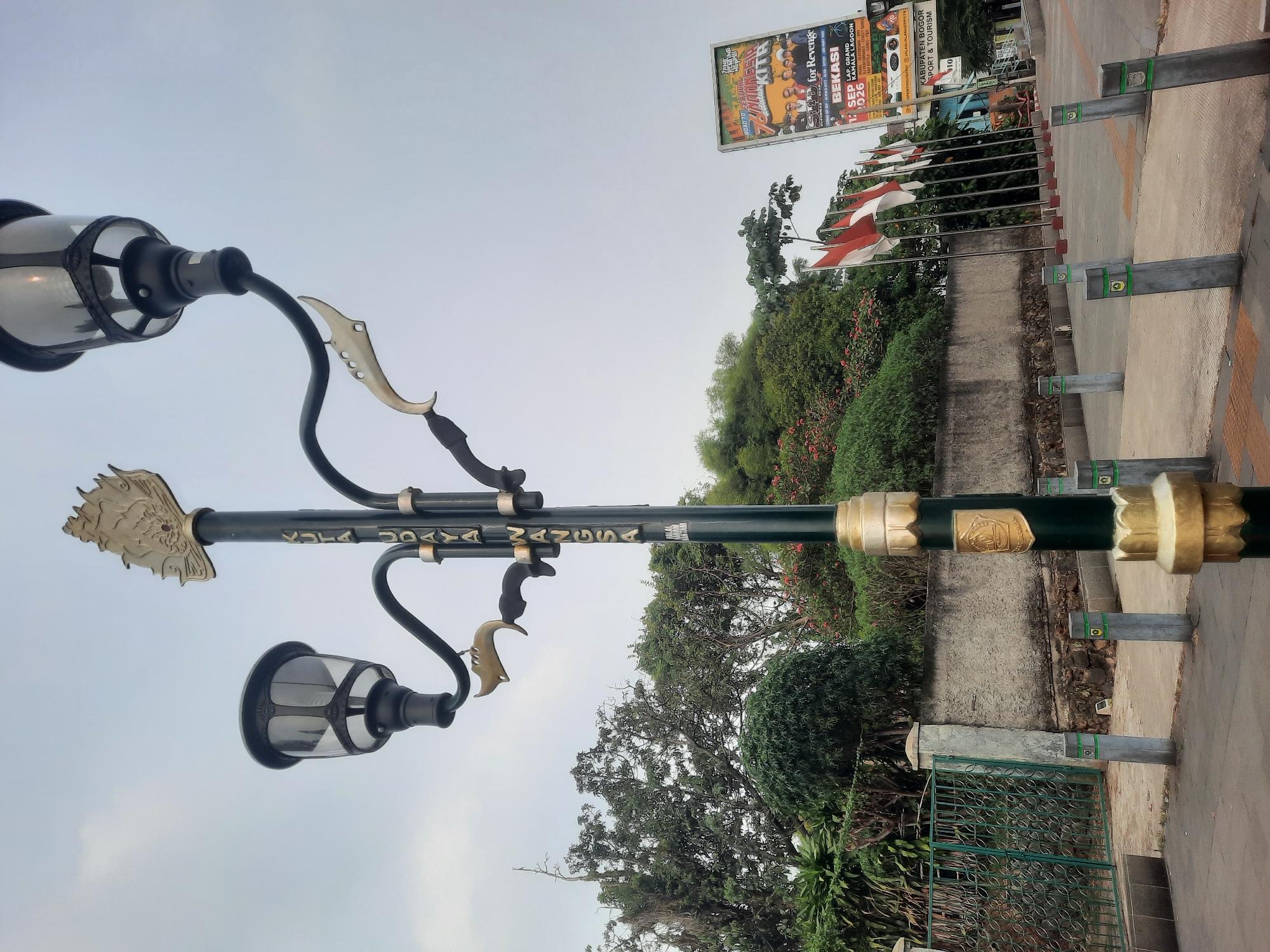The image shows a street scene in Sukaraja, Indonesia, featuring a tall, ornate lamppost with multiple lights and decorative elements. The lamppost is dark green with gold accents, including a crest at its base and stylized figures along the pole. The text "KOTA UDAYA WANGSA" is visible vertically on the lamppost in gold lettering. To the left of the lamppost, a green metal fence with a decorative pattern encloses an area with lush green trees and bushes. A grey concrete wall runs behind the fence and extends to the right.
In the background, a row of Indonesian flags on poles is displayed. To the right of the flags, a large billboard advertising a "76 INDONESIA KITA" event and a "BEKASI" event is prominently visible. Several concrete bollards with green accents are lined up along the pavement in front of the wall. The sky is overcast, suggesting a daytime scene. The ground is paved with concrete and includes some textured tilework. No people are visible in the image.