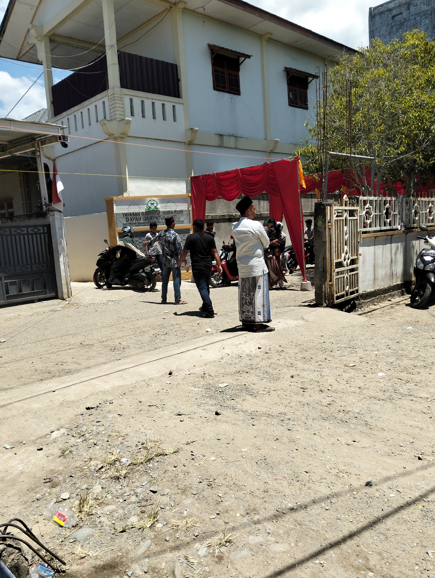 The image depicts an outdoor scene in Teungoh, Indonesia, likely in front of a building belonging to the "YAYASAN QARYATUL HUDA" organization. The building is white with a balcony supported by columns. A red curtain is draped across the front of the building, suggesting an event or gathering.
In the foreground, the ground is unpaved and appears to be dirt and gravel, with some scattered debris. A few people are visible in the middle ground. One man, wearing a white shirt and a sarong, stands facing away from the camera. To his left, two men in casual attire are standing near a motorcycle. Another man on a motorcycle, wearing a helmet, is also present. Further back, near an open gate, more individuals are partially visible. A white motorcycle is parked on the right side of the image. The environment appears to be a street or pathway leading to the building, with a fence visible on the right. The sky is partly cloudy.