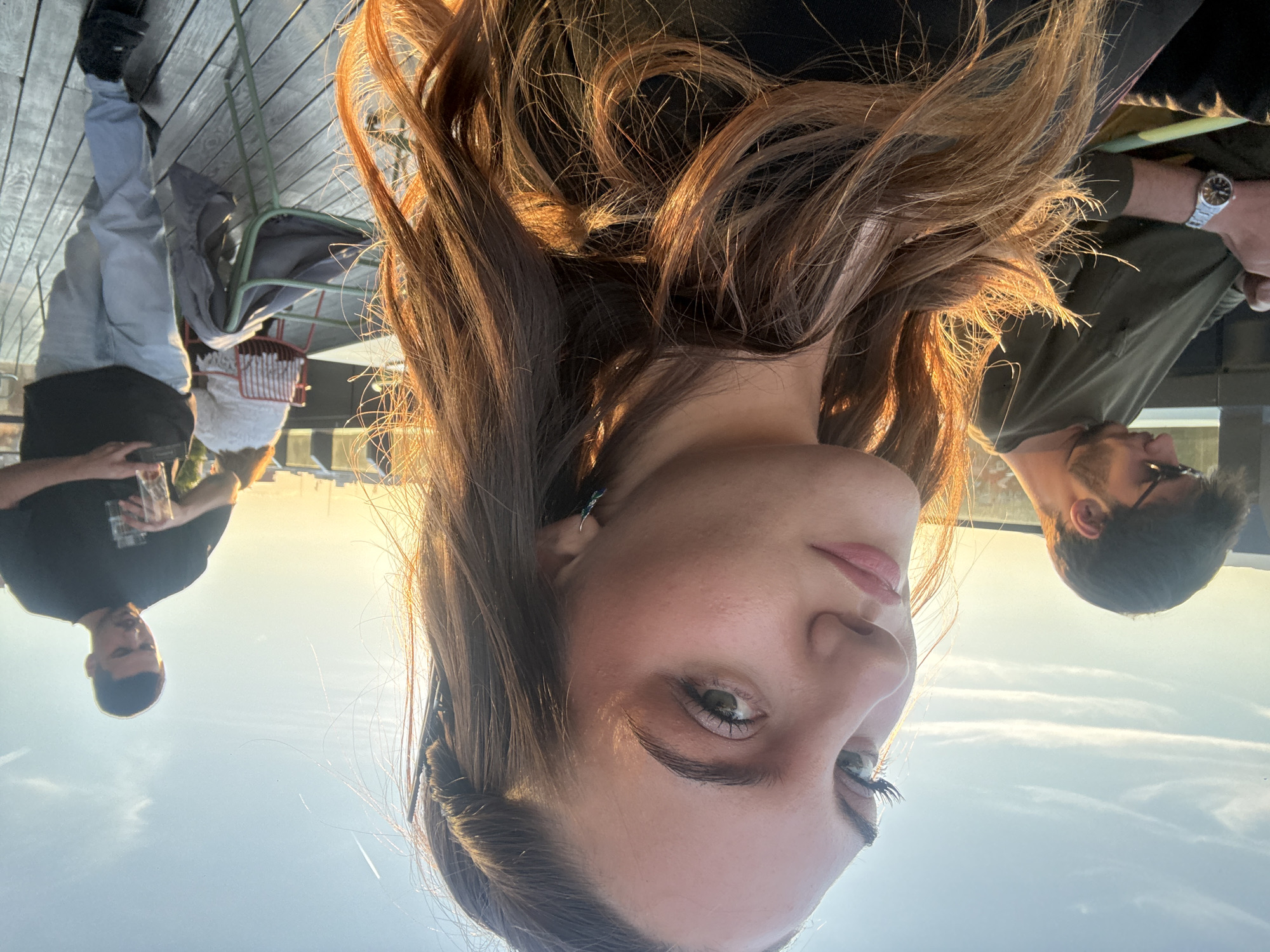 The image is a selfie taken from an upside-down perspective. The subject is a woman with light skin, brown hair, and green eyes. She is wearing dark clothing. Behind her and in the background are three men. One man is wearing a black shirt and holding a glass; another is wearing a green shirt and a watch. The third man is partially visible behind the woman's hair. The setting appears to be an outdoor patio or rooftop. The foreground appears to be a wooden deck. The scene is lit by natural light. The location is the city of Sofia, Bulgaria.