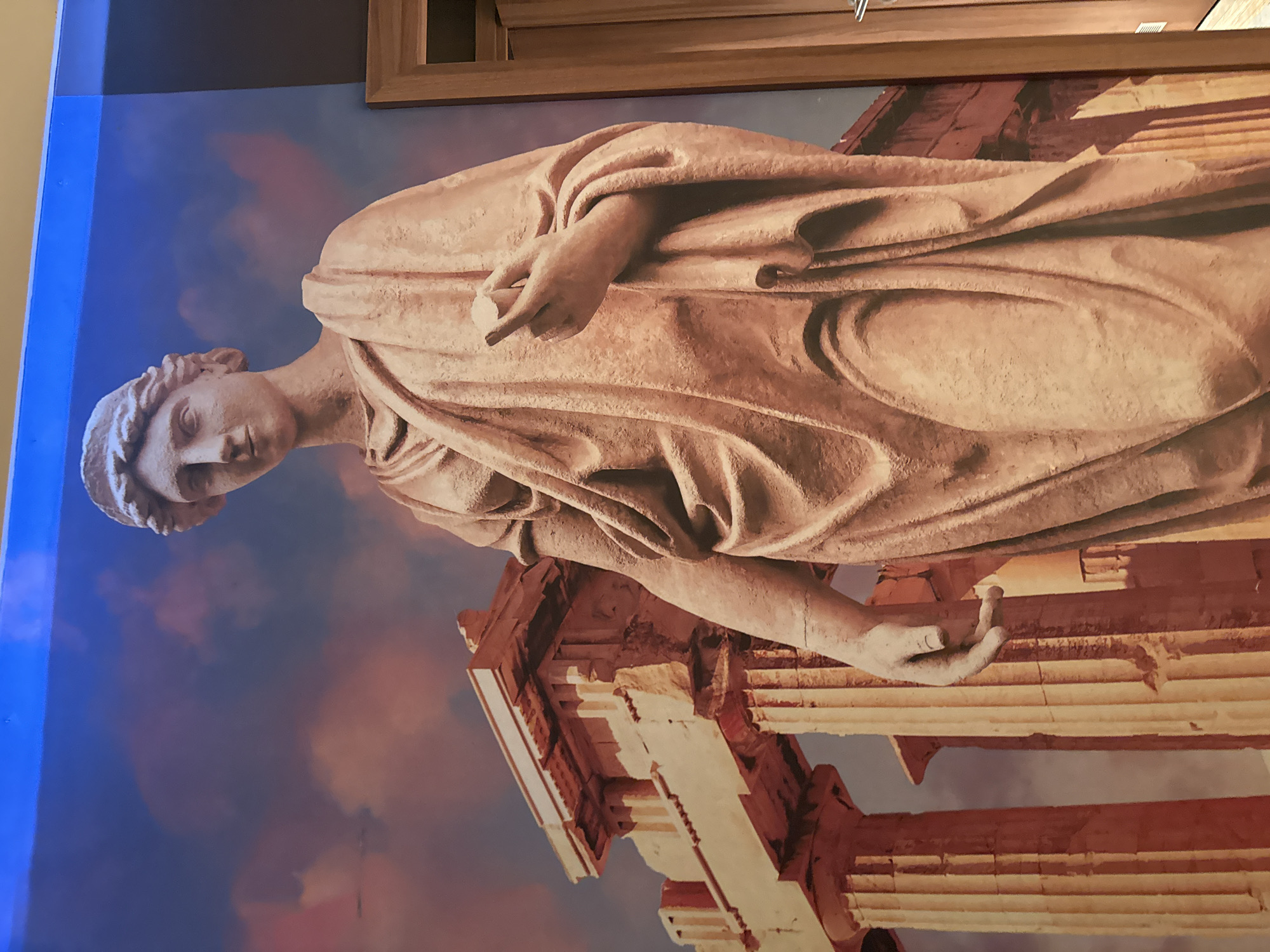 The image shows a mural of a Greco-Roman statue with a classical architectural element in the background, possibly the Parthenon in Athens. The mural is on a wall of the city of Burgas, Bulgaria, or a representation thereof. The statue is draped in a classical toga. The background is a painted blue and red sky. The mural is framed at the top by a light brown wooden panel. The image is rotated 90 degrees counter-clockwise.