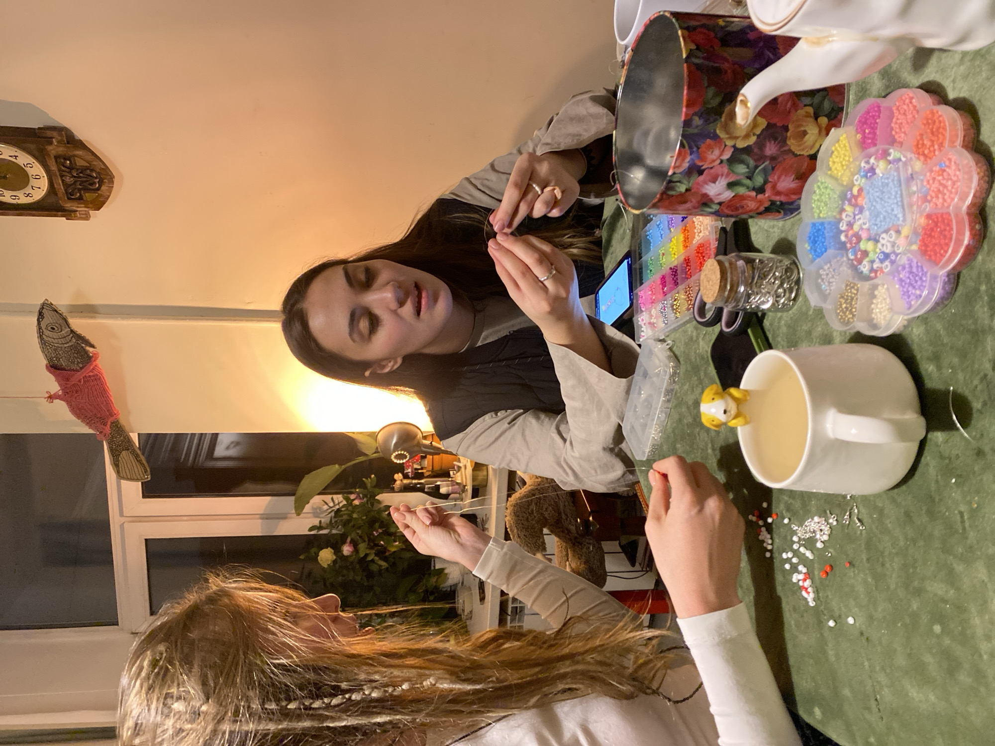 Two women are sitting at a table, crafting beaded jewelry. One woman has dark hair and is wearing a dark vest over a light shirt. She is holding a piece of jewelry and looking at it. The second woman has long, blonde hair with braids, wearing a white long-sleeved shirt. She is also holding a piece of jewelry. There is a green tablecloth on the table, and a variety of beads, containers, and crafting supplies are scattered around. There is a mug on the table with a small yellow figurine attached. A floral-patterned tin can and a teapot are also visible on the table. In the background, there is a window, a lamp, and a plant. The woman is next to an item that appears to be a pink fish-shaped ornament hanging on the wall. There is also a clock on the wall. It is nighttime, given the lights and the darkened window. There is no information that can infer that the image was taken in Moscow, Russia.