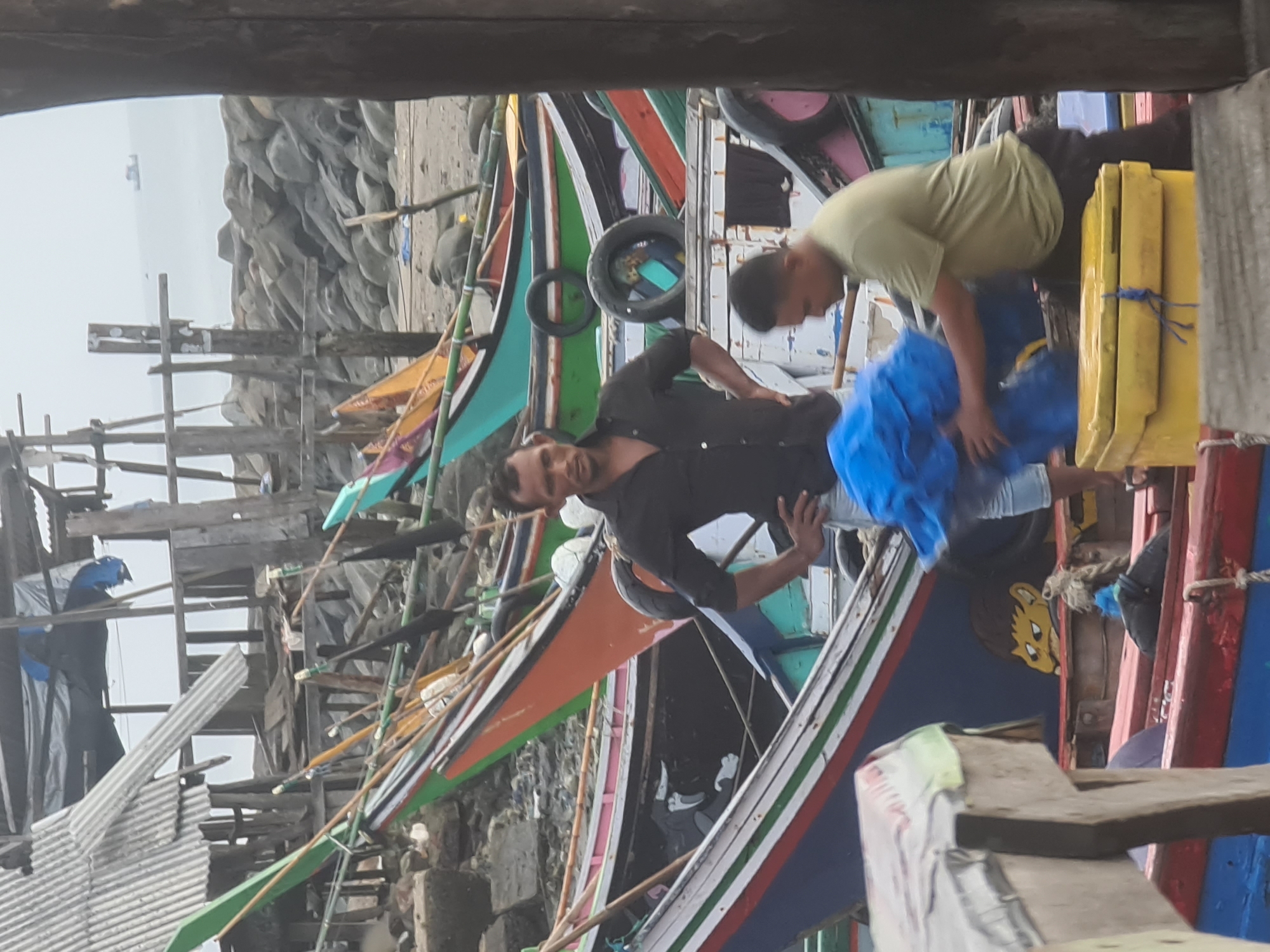 The image depicts a scene in Lhokseumawe, Indonesia, likely near a harbor or fishing area, with numerous colorful fishing boats docked. Two men are actively engaged in work. In the foreground, a man in a black shirt and jeans stands with his hands on his hips, looking towards the viewer. To his right, another man in a light green t-shirt and dark pants is kneeling, carrying a large, bright blue tarp bundle. A yellow cooler is positioned in front of him. The boats are varied in color, with orange, green, pink, and blue being prominent. Some boats have decorative paintings on their hulls. The background reveals a rocky shoreline, the ocean, and a slightly overcast sky. Structures made of wood and corrugated metal are visible behind the boats, suggesting a processing or storage area. The overall impression is one of a working maritime environment.