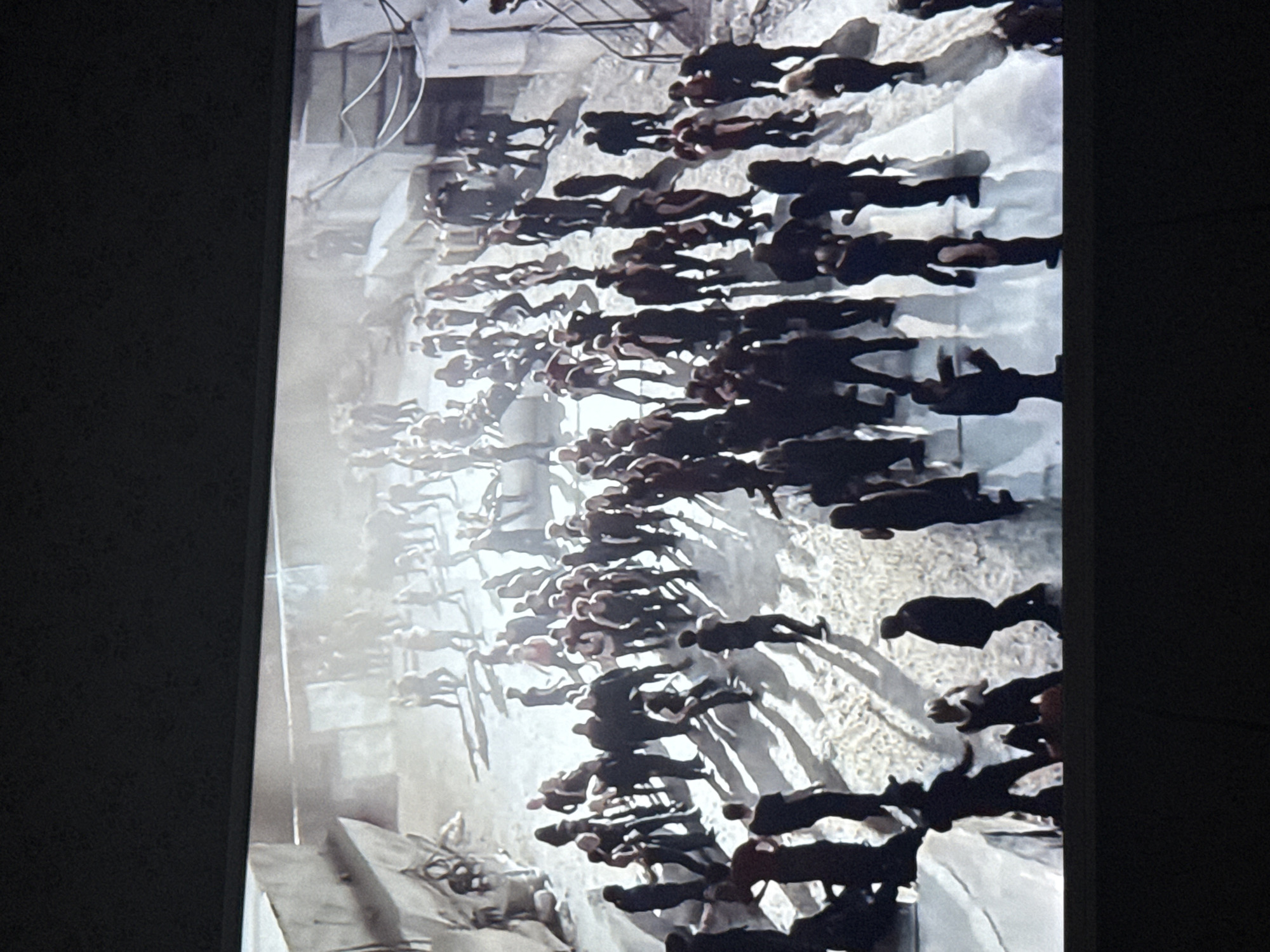 A high-angle shot displays a crowd of people, primarily silhouetted against a bright, concrete-laden background. The environment appears urban, potentially a plaza or public space with visible architectural structures and features. The people are tightly packed and oriented in a similar direction. The scene is generally monochromatic with dark shadows, suggestive of a sunny day. The location in Mytishchi, Russia cannot be determined from the image.