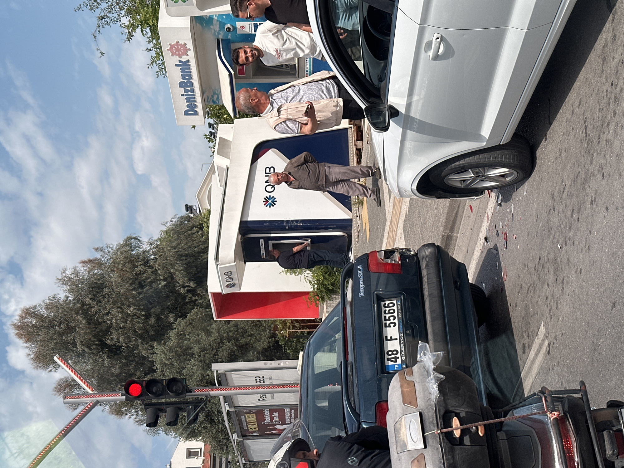 The image depicts a street scene in Fethiye, Turkey, featuring several people around ATMs, a white car, and traffic stopped at a red light. There are two ATMs, one branded "DenizBank" and the other with "QNB" logos. Three men are standing near the DenizBank ATM, seemingly engaged in conversation. A man is using the QNB ATM. A white car is parked nearby, with its front partially visible. Behind the car, traffic is stopped at a red traffic light. A motorcycle is in front of the camera's point of view. A blue car with the license plate "48 F 5566" is also visible. Debris is scattered on the road next to the white car. Trees are on the left.