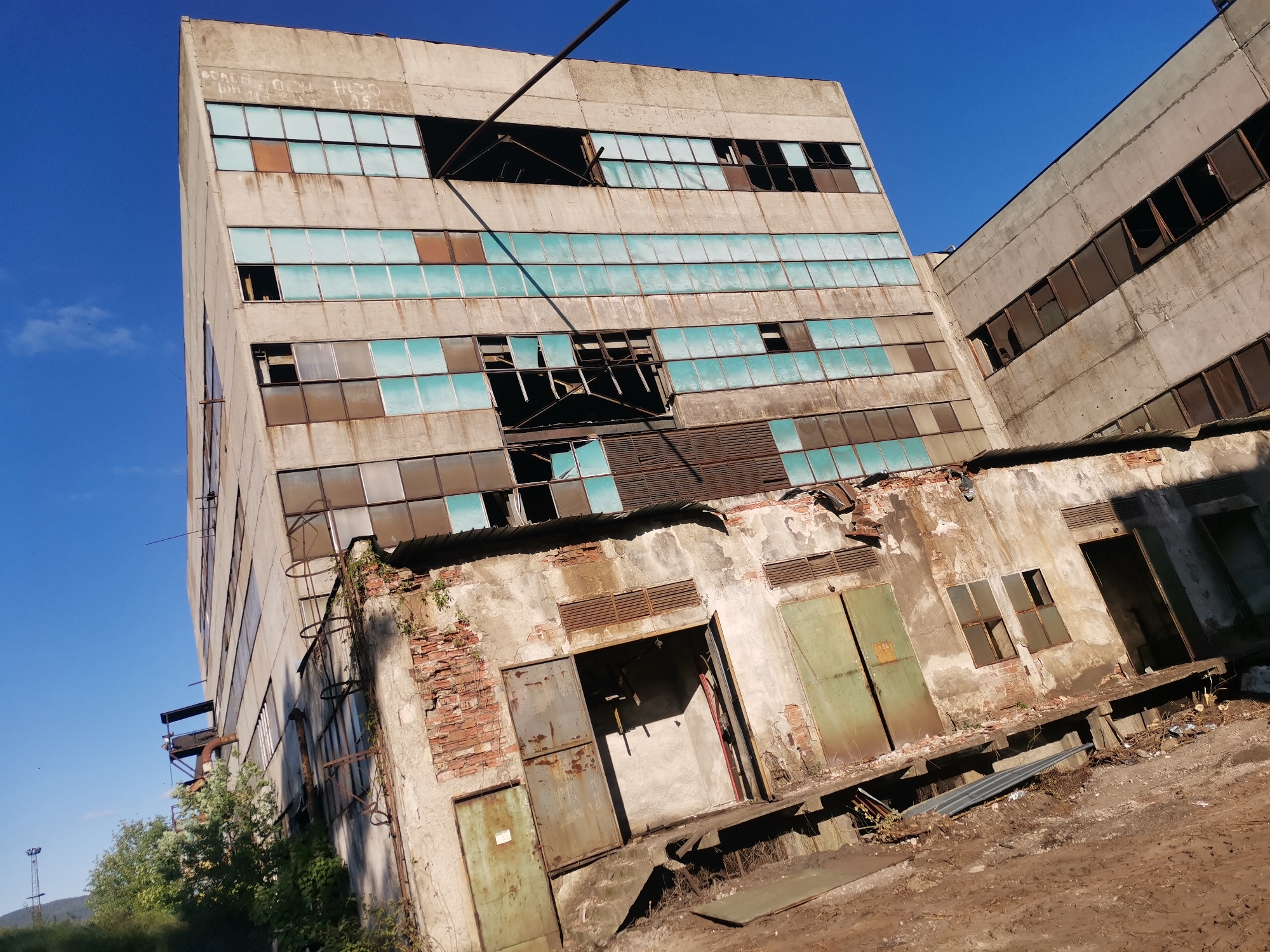 The image depicts an abandoned industrial building complex located in Zhivkovo, Bulgaria. The buildings are primarily constructed of weathered concrete and have numerous rows of windows, many of which are broken or missing. The windows that remain show a blue-green tint. The facade of one building has visible brickwork and large metal doors, suggesting it was used for storage or loading. The surrounding ground is bare and uneven. The overall impression is one of decay and disuse. A clear blue sky is visible in the background.