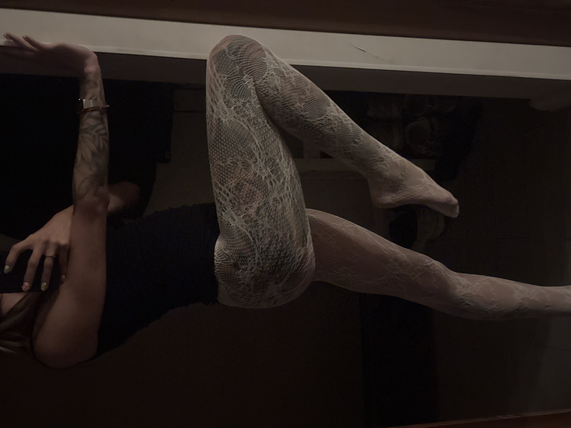 A person is standing in a dark room, leaning against a white door frame. They are wearing a black tank top and white lace patterned tights. Their left leg is bent at the knee and their foot is lifted off the ground. Their right arm is resting on the door frame, and they are holding a phone in their left hand. A tattoo is visible on their right arm. The environment is dimly lit, with some furniture visible in the background. It is not possible to determine the location in Moscow, Russia from the image.