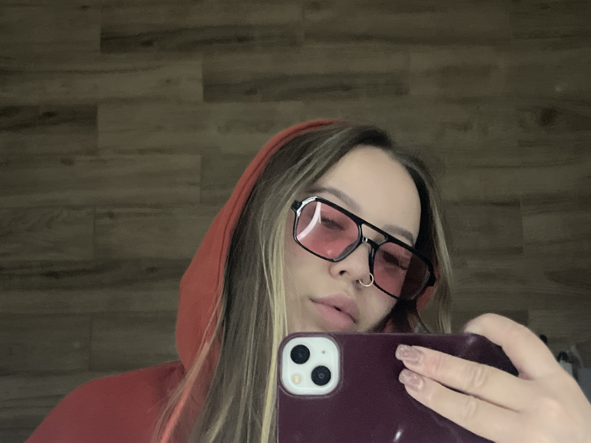 An individual is captured in a close-up, waist-up portrait, seemingly taking a selfie indoors in Krasnodar, Russia. The subject, a person with long, light brown hair featuring blonde highlights, is wearing a red or coral-colored hooded sweatshirt with the hood up. They are looking down and slightly to their right.
A prominent feature is the pair of large, black-framed glasses with distinctive pink-tinted lenses that obscure the eyes. A small, silver hoop nose ring is visible on their septum. Their lips are slightly parted, revealing no particular expression.
The subject holds a dark purple or burgundy smartphone case, displaying the dual camera lens system (two large circular lenses and a smaller flash) of what appears to be an iPhone model. Their hand, adorned with light-colored or glittery rectangular-shaped fingernails, is visible gripping the phone.
The background consists of a light brown wooden wall with horizontal planks, suggesting an indoor setting like a room or hallway. The lighting is soft and even, without harsh shadows. No text is visible in the image.