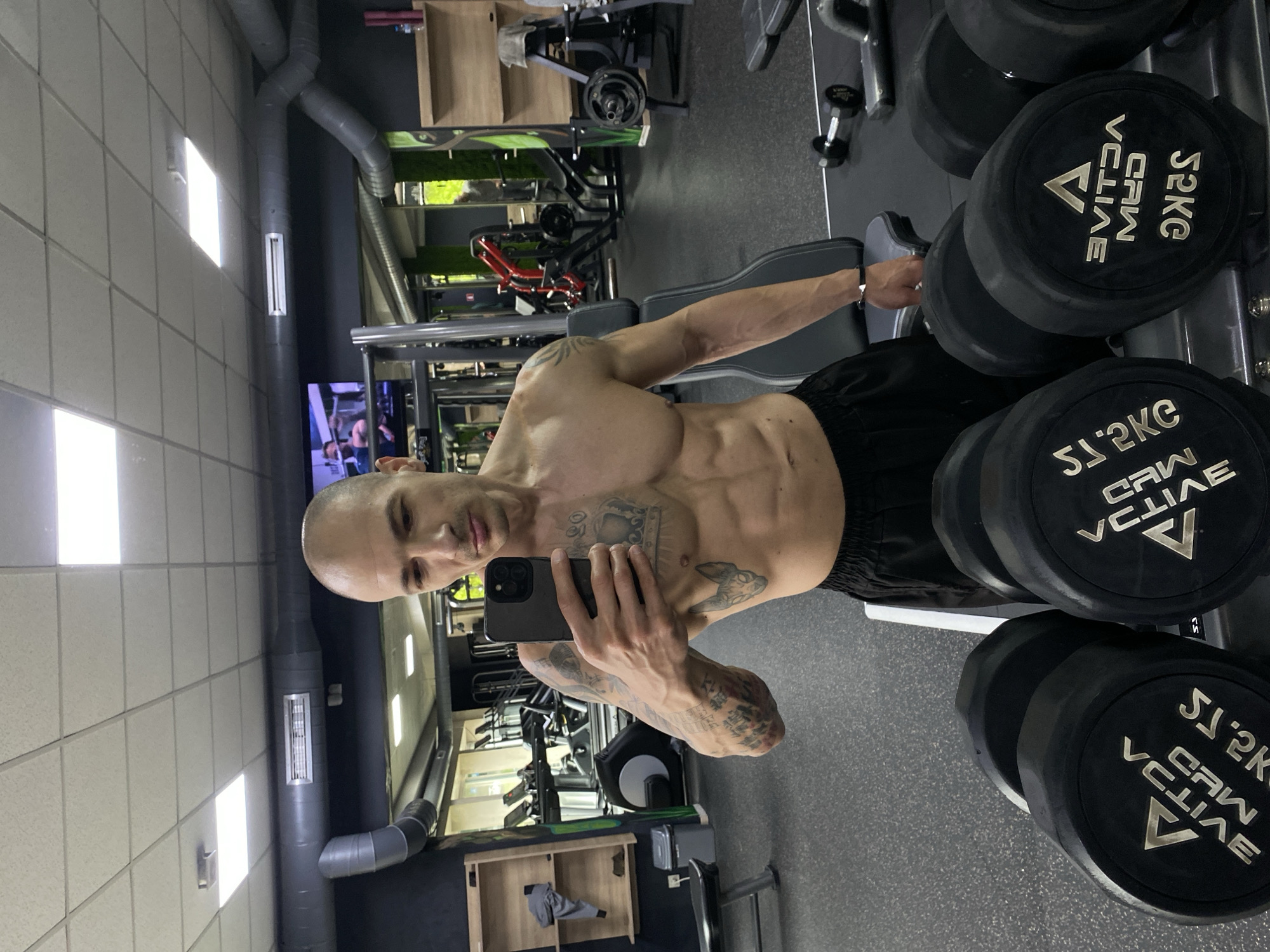 A muscular, shirtless man stands in a gym, facing the camera and holding a smartphone in his left hand, presumably taking a selfie. The gym has various exercise machines and a rack of dumbbells behind him. The weights are labeled "52 KG VCLIVE", which indicates the gym's branding. The background features gray walls, a drop ceiling with fluorescent lighting, and ventilation ducts. Based on the "VCLIVE" branding on the weights, the location is likely Vclive Fitness Center in Pleven, Bulgaria.