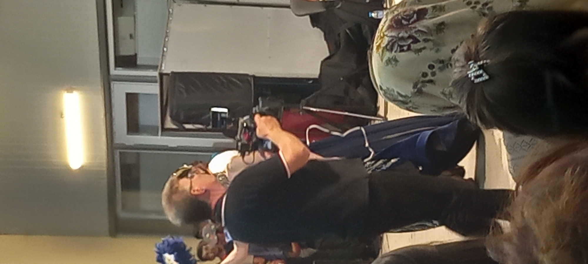 The image depicts an outdoor scene, possibly in Lyuben, Bulgaria, with several individuals present. In the foreground, a man wearing glasses and a black polo shirt with white trim is holding a video camera. Behind him, a person in a blue garment with white trim is visible. Further back, a group of people are gathered, some with faces obscured. The background shows a building with large windows and a fluorescent light fixture overhead. There are also large, dark objects that appear to be equipment or bags. The overall environment suggests an event or gathering where video recording is taking place.