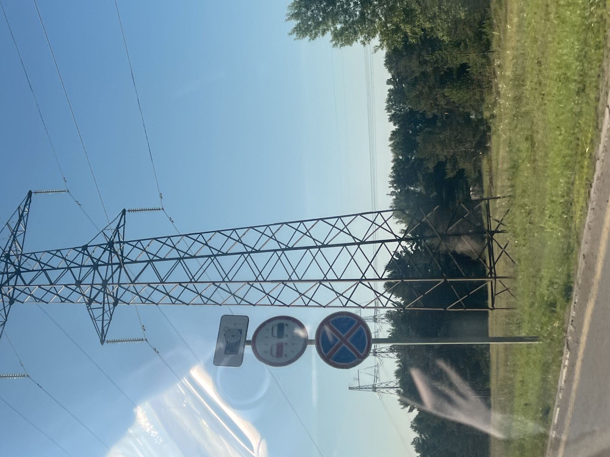 This image captures a rural roadside scene dominated by a large metal power transmission tower under a clear blue sky. Several wires are strung between towers, indicating a power line. To the left of the main tower, a pole displays three traffic signs stacked vertically. The top sign is rectangular with an image of a camera and a flower. Below it, a circular red-bordered sign depicts two cars, one red and one black, with a diagonal red line, suggesting a prohibition on passing or parking related to vehicles. The bottom sign is circular with a blue background and a red cross, indicating "no parking" or "no stopping." In the background, a dense forest borders a grassy field. The perspective suggests the photo was taken from a moving vehicle, as indicated by a blurred, ghostly white shape on the left side of the image, likely a reflection or smudge on the window. The ground in the foreground is a paved road with a grassy shoulder. There are no people visible in the frame. The location is identified as Noviy, Russia.
