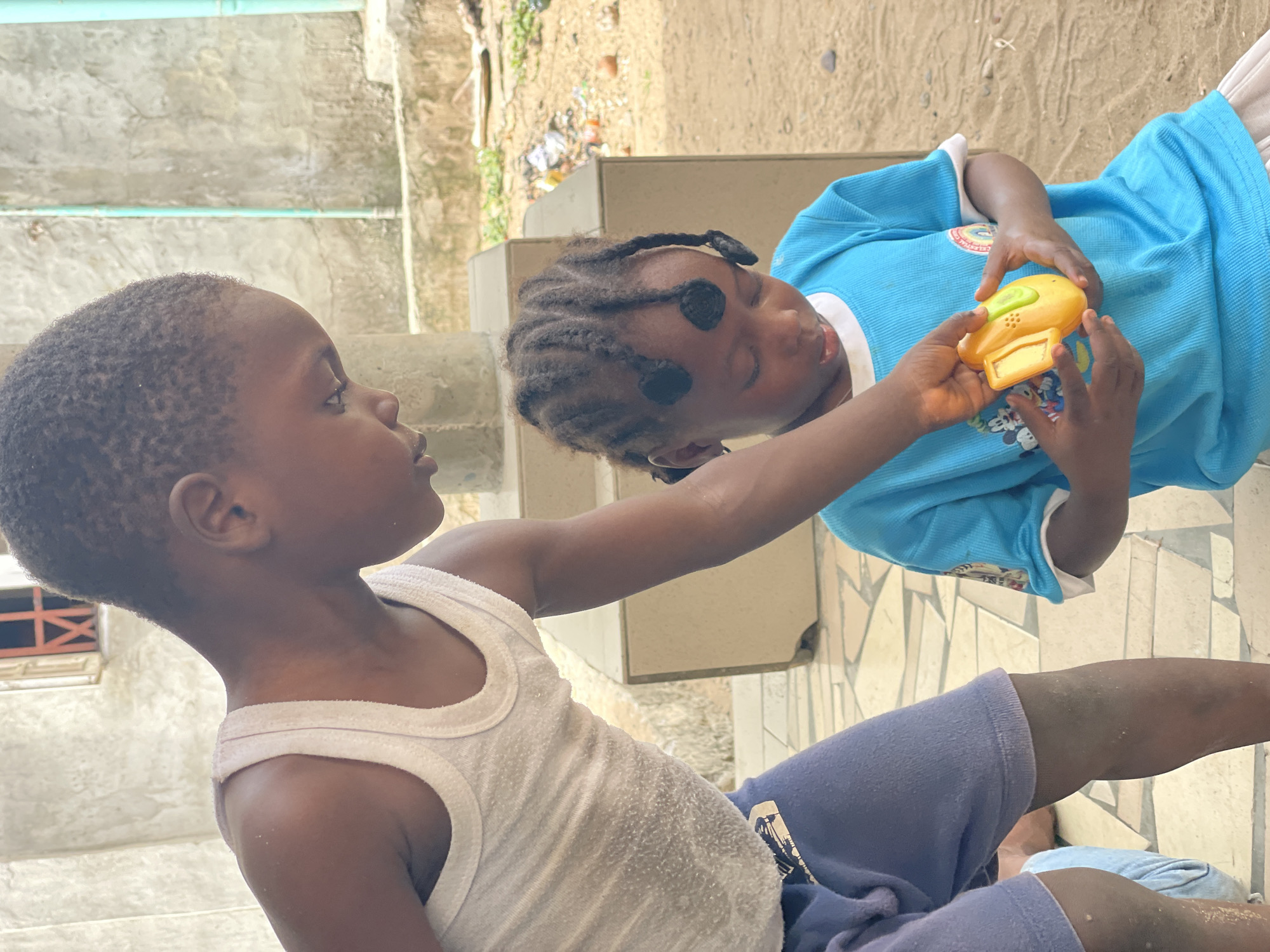 Two young children are interacting in an outdoor setting. One child, wearing a white tank top and blue shorts, is extending their arm toward the other child. The second child is wearing a light blue shirt and is holding a yellow toy with both hands. The background shows a building with concrete steps, tiled ground, and some vegetation. The location is Badagry, Nigeria.