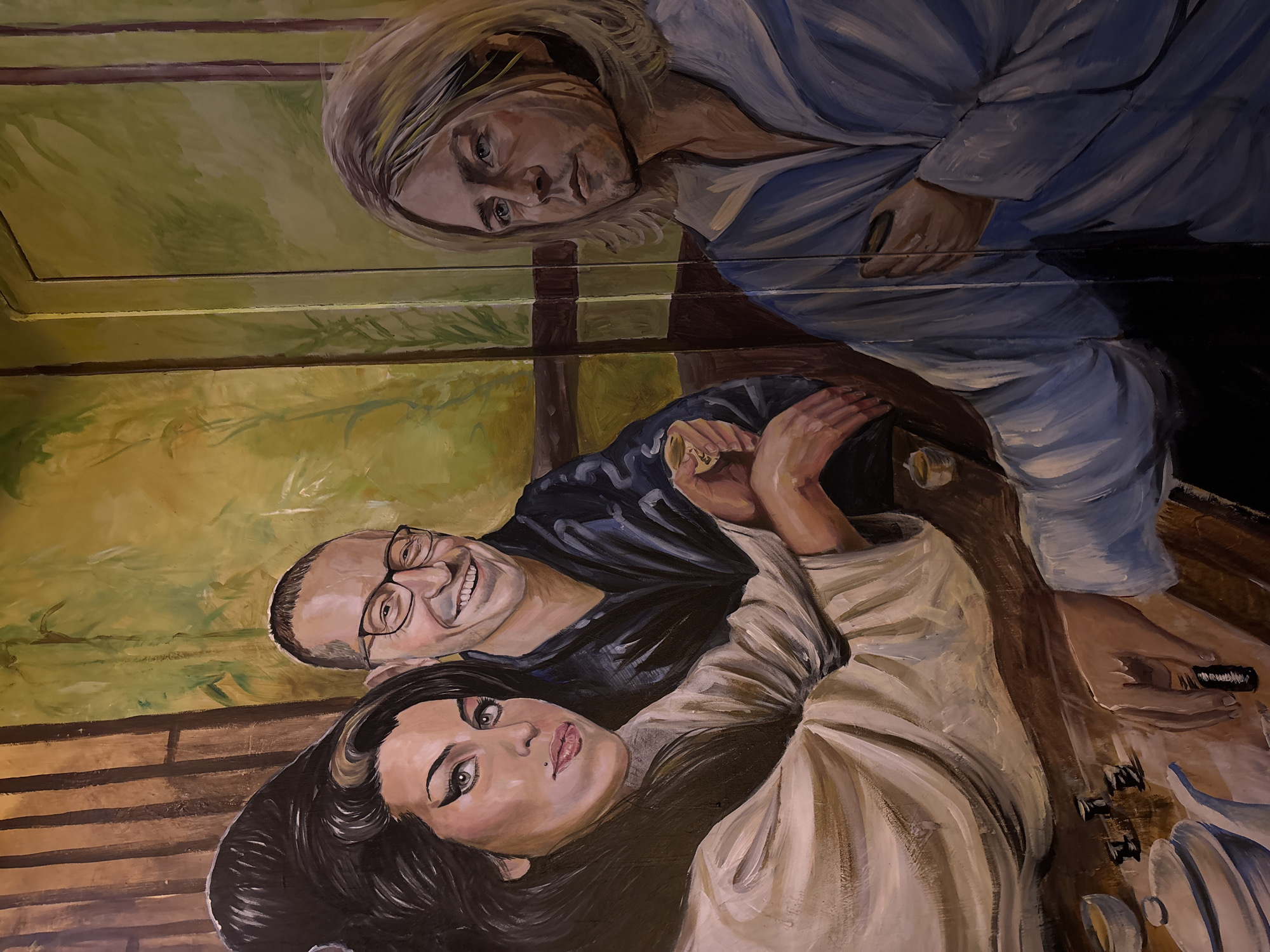 The image is a mural on a wall at the bar, "The Bar Next Door," located in North Kuta, Indonesia. The mural depicts three people: Kurt Cobain, Amy Winehouse, and Walter Becker. Kurt Cobain, with long blond hair, wears a blue shirt and looks downward. Amy Winehouse has black hair with a mole on her chin. Walter Becker has glasses and a shaved head and is smiling. Winehouse and Becker are positioned at a table with a guitar.