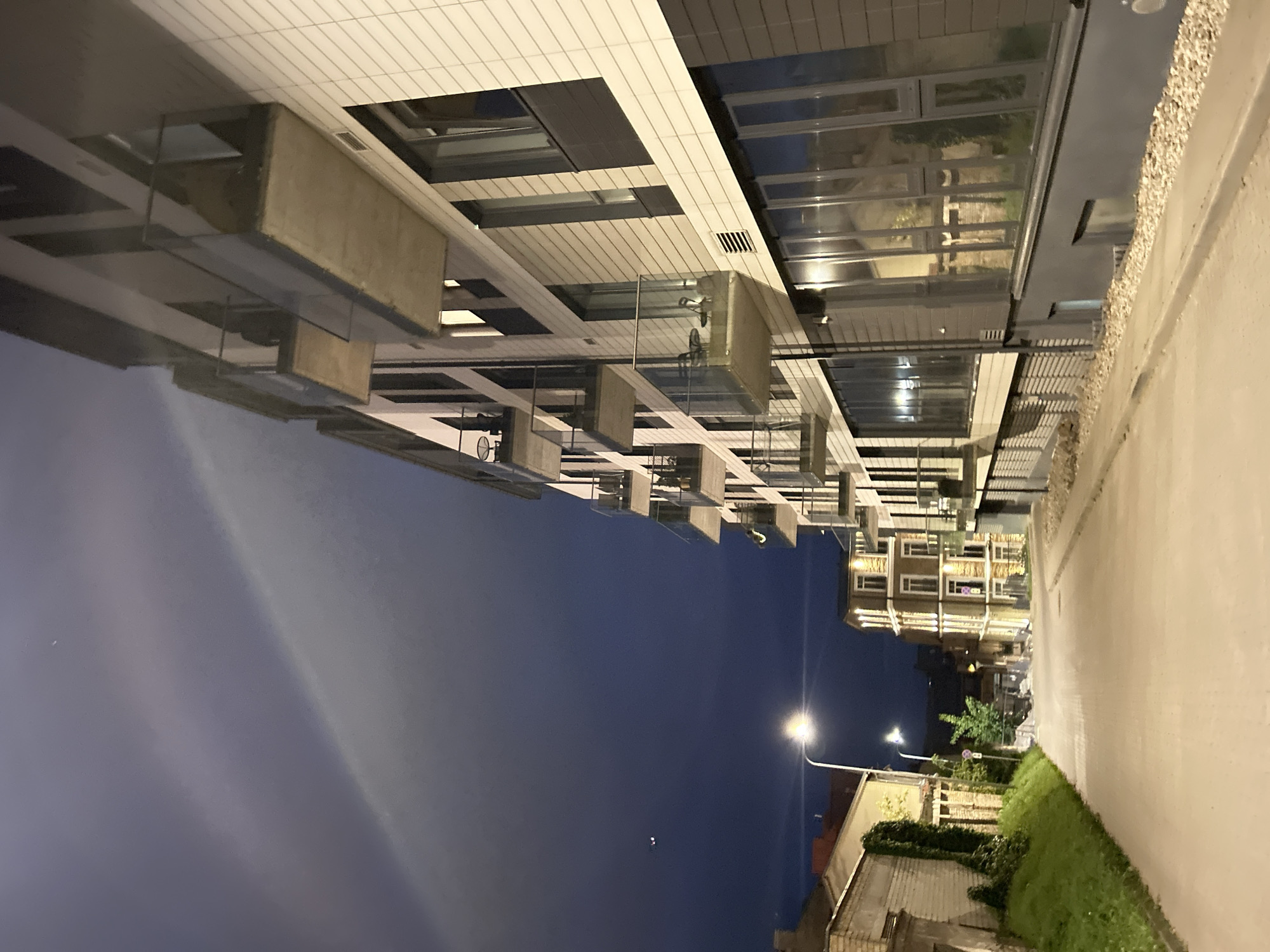 The image depicts a street scene at night in Vilnius, Lithuania. A modern apartment building with balconies dominates the right side of the frame, its facade clad in light-colored tiles. To the left, older brick buildings and a green slope are visible. A street lamp illuminates the asphalt road, which slopes gently uphill. The sky is dark blue, with some atmospheric haze or clouds creating a gradient of light towards the top left corner. A small object, possibly an aircraft, is faintly visible in the upper left quadrant of the sky. There are no people or discernible activities in the scene.