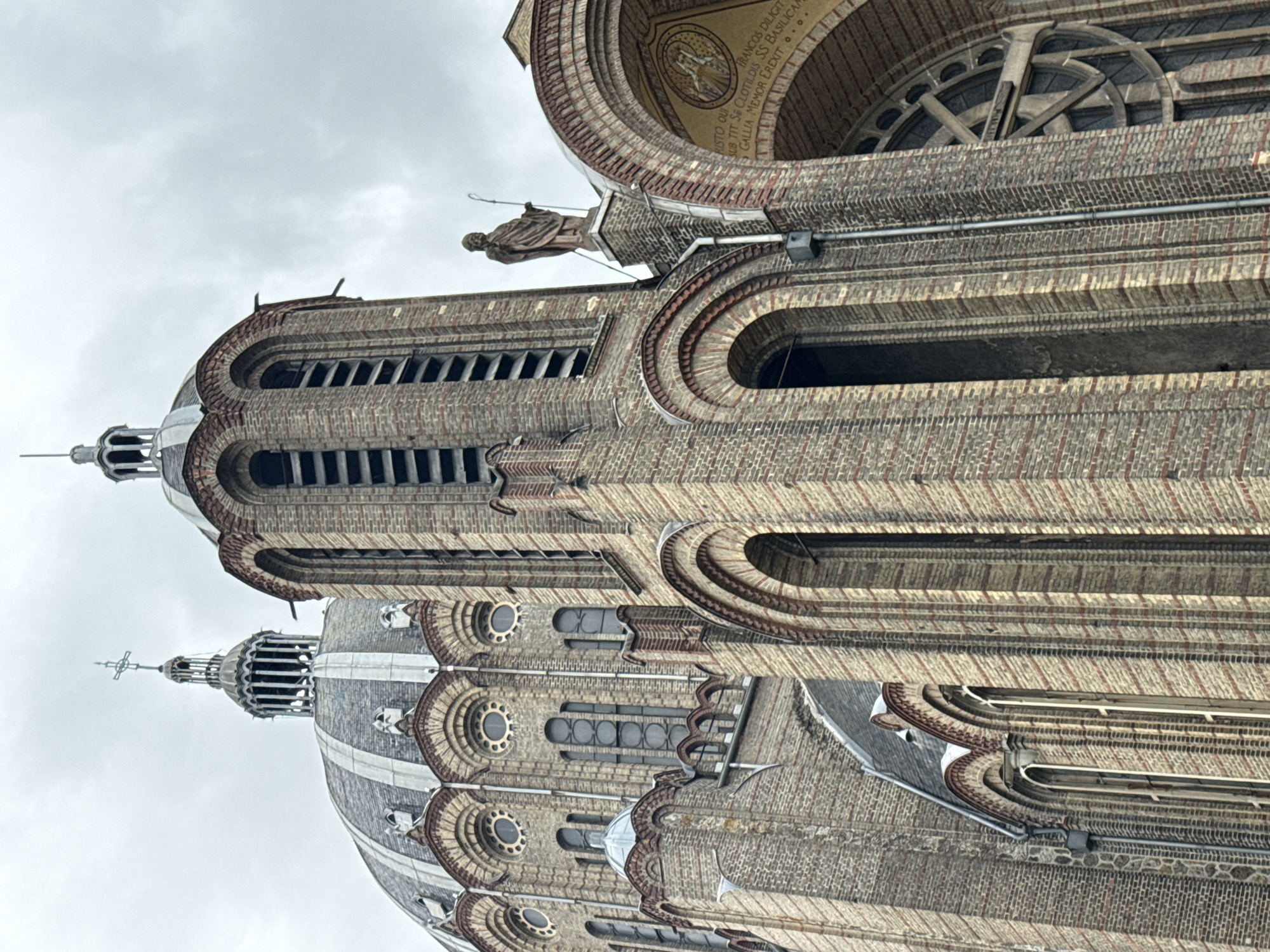 This image presents a grand, intricately designed basilica in Reims, France, captured from a low, upward-looking perspective on an overcast day. The building, likely the Basilica of Saint-Remi, showcases a distinct architectural style characterized by extensive multi-toned brickwork, numerous arched windows, and decorative elements.
Two prominent sections with domes and spires dominate the vertical composition. The closer section features a tall, narrow tower with louvered openings, topped by a gray dome and a slender spire adorned with a cross. A robed statue, possibly a saint, stands on a ledge below the dome. Beneath the statue, a large semi-circular arch frames a detailed mosaic depicting a central figure and partial Latin inscriptions. Visible text from this mosaic includes "RISTO QUI", "FRANCOS DILIGIT", "SUB TIT SS CLOTILDIS SS", "GALLIA MEMOR EREXIT", and "BASILICAN", suggesting dedications like "Christ who loves the Franks, under the title of Saints Clotilde, Gaul remembering erected this Basilica."
Below the mosaic, a large rose window with radial spokes adds another layer of detail. The building's walls are textured with varying brick patterns and recessed arches, creating a sense of depth and ornamentation. Numerous smaller circular and arched windows are visible across the structure. The cloudy sky provides a soft, even light, emphasizing the architectural details without harsh shadows. No human activity is visible, focusing solely on the majestic edifice.
