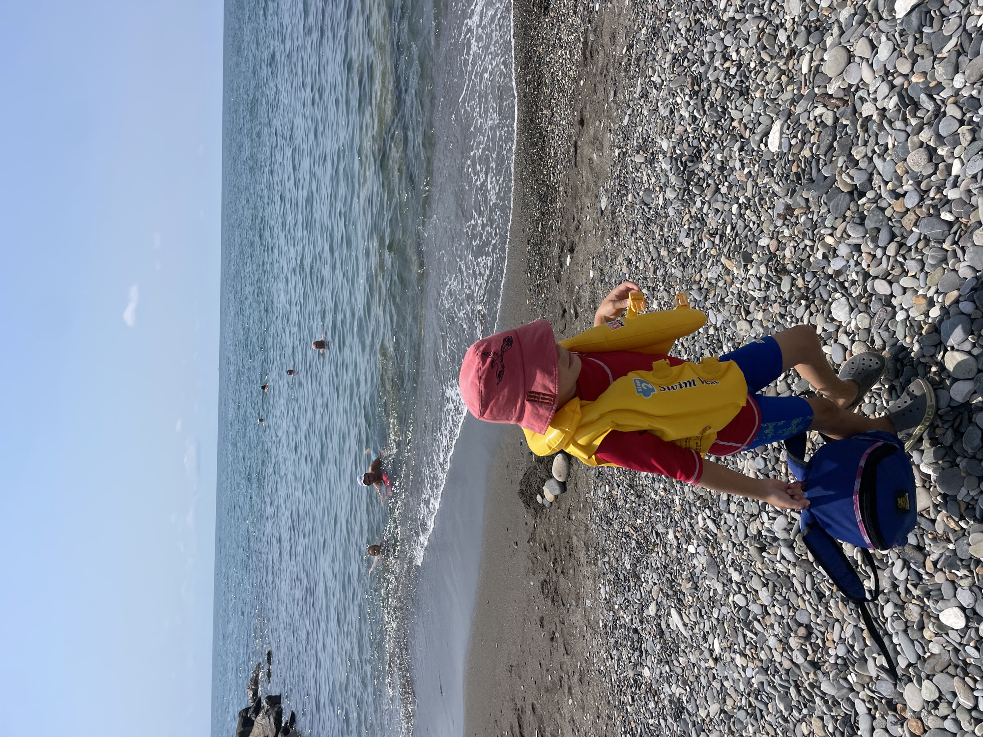 A young child wearing a red hat and a yellow life vest walks on a pebble beach, carrying a blue backpack in their left hand. The child is wearing blue swim trunks and gray Crocs-style shoes. The child is looking down and holding a yellow object in their right hand. In the background, the sea is calm with small waves and several people are swimming. The sky is clear and blue. The beach is covered in smooth, gray pebbles of various sizes. The location is likely a beach in Sochi, Russia, given the stony shore and the presence of people swimming in the sea on a sunny day.