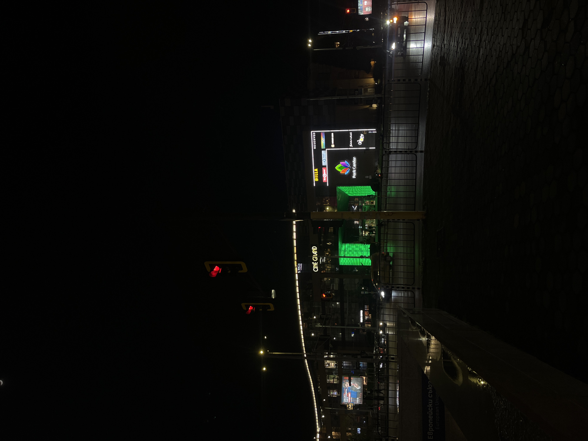 The image depicts an urban night scene in Sofia, Bulgaria, showcasing illuminated commercial buildings and city infrastructure. The view is dominated by a multi-story complex, likely a shopping mall or entertainment center, brightly lit against the dark sky. Prominent signs on the building include 'Park Center' with a colorful logo, 'BILLA' (a supermarket), 'Reserved' (a fashion retailer), 'Technopolis', 'Subway', 'Graffiti', and 'Cine Grand' (a cinema). A small billboard featuring people is also visible near the 'Cine Grand' sign.
A distinctive section of the building or an architectural feature glows with a vibrant green light. In the foreground, a paved area with hexagonal block patterns is visible, separated from the building by metal barriers or fences. Two red traffic lights are prominently displayed on poles in the mid-ground. Faint lights from vehicles can be seen, suggesting ongoing traffic, though no individual vehicles or people are clearly distinguishable. A slender crescent moon is faintly visible in the dark sky on the left. On a metallic structure in the lower right foreground, text in both Bulgarian and English reads 'Европейски съюз' and 'European Union', providing a clear geographical identifier. The scene conveys a calm atmosphere typical of a clear night in a modern city.