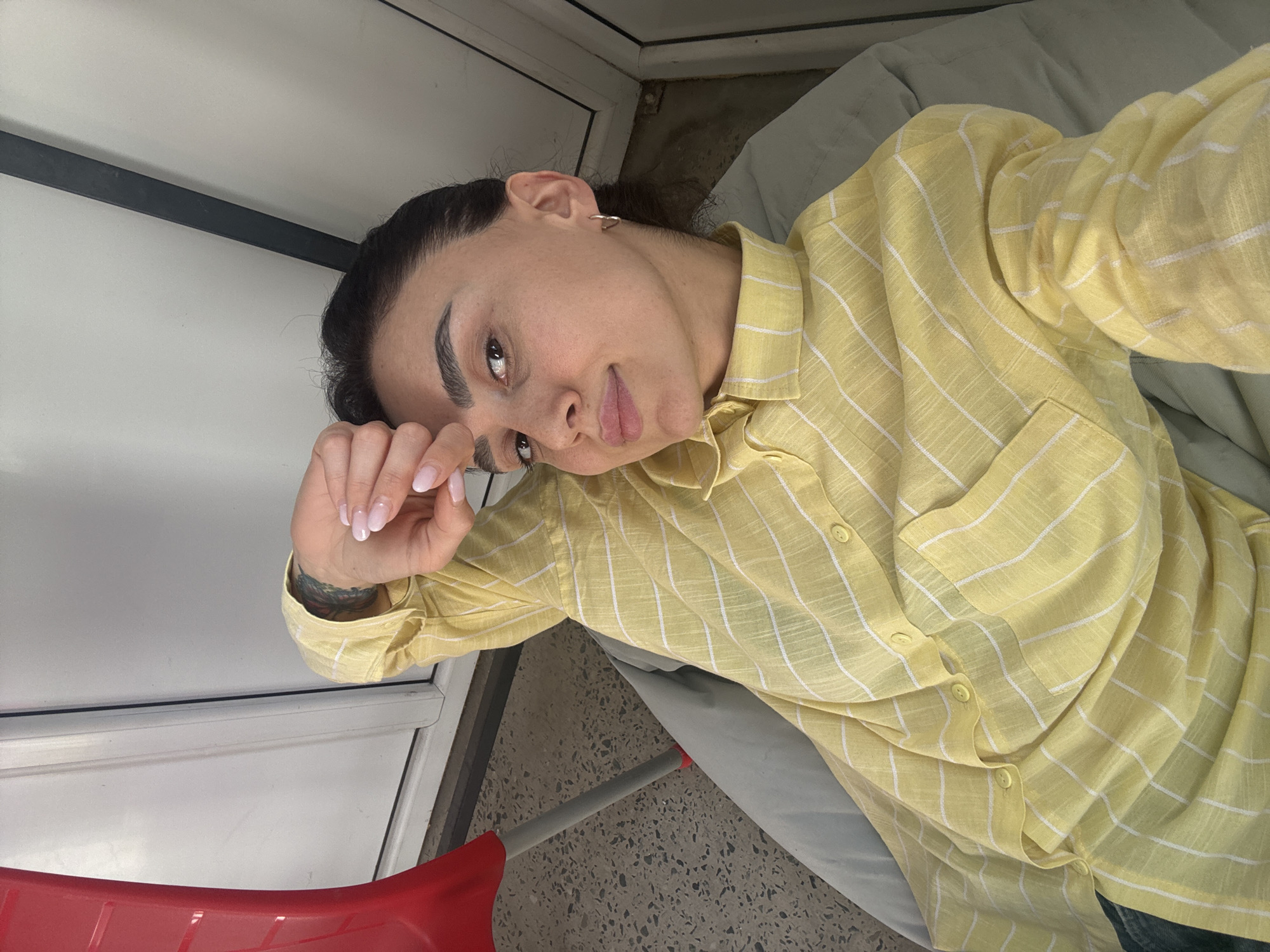 A woman with dark hair pulled back is lying on a light green surface, likely a bed, wearing a yellow and white striped button-down shirt. Her left hand is near her face, and she's looking at the camera. She has a small hoop earring in her left ear. There's a tattoo visible on her left wrist. The background includes a white wall with a dark trim, a red plastic object (possibly part of a chair or cleaning tool) and a floor with small dark spots. The picture appears to be taken indoors. The city of Tbilisi, Georgia, cannot be confirmed based on this image alone.