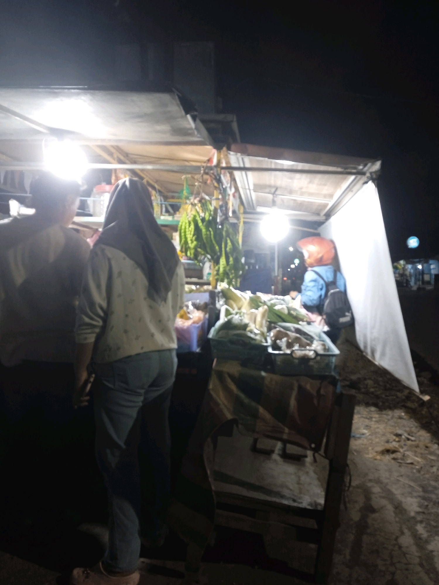 The image depicts an outdoor scene at night, likely a street food stall in Langensari, Indonesia. Two individuals are visible. In the foreground and to the left, a person wearing a light-colored shirt is partially visible from behind, facing away from the camera. Next to them, a person wearing a grey hijab and a light-colored patterned sweater is also facing away. They are wearing blue jeans and light-colored shoes. Behind them, a stall is set up with a variety of items, including what appears to be fresh produce such as corn, displayed in plastic containers. The stall is illuminated by bright overhead lights. Further back, a person wearing a blue jacket and an orange helmet with a black backpack is standing near the stall. The background shows a dimly lit street with some distant lights, suggesting an urban or semi-urban environment. The ground appears to be dirt or gravel. The overall atmosphere is that of a typical night market or roadside vendor.